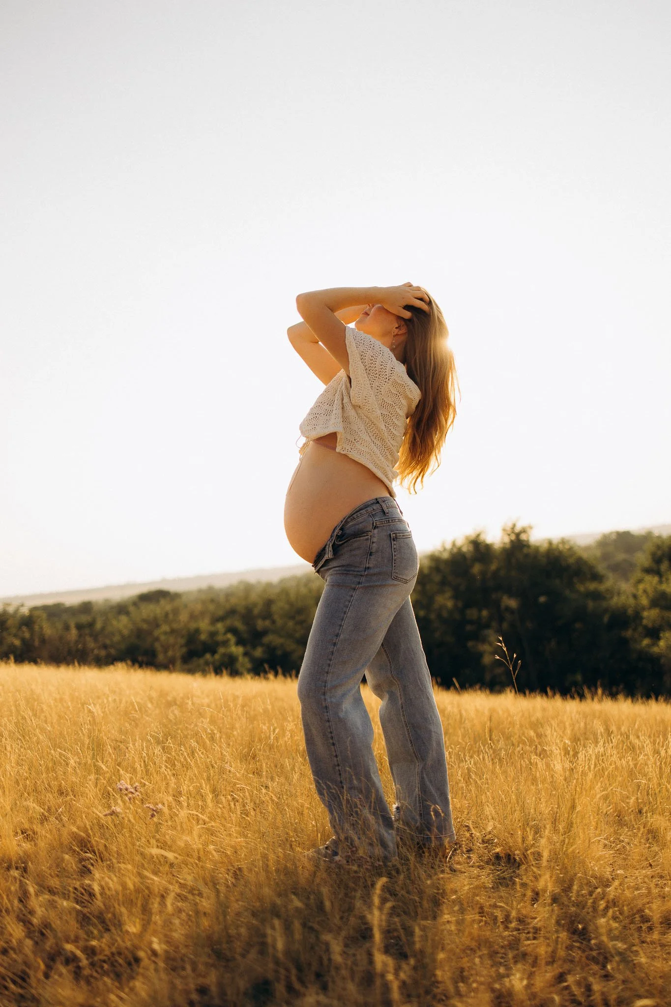 A pregnant woman standing in a field of tall, golden grass during sunset, with one hand on her head and the other on her belly, wearing a white crochet top and jeans.