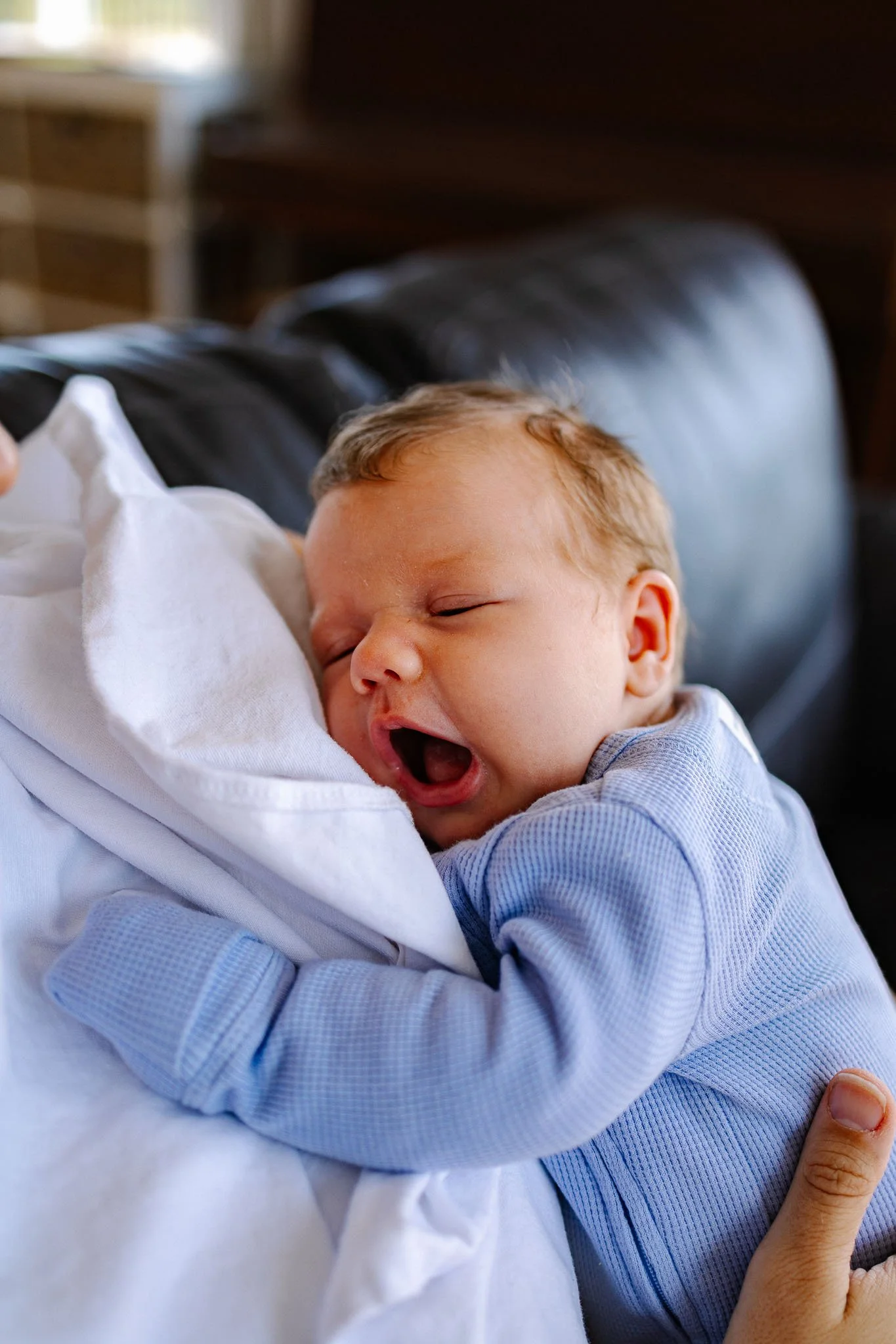 Close-up of a newborn baby yawning and sleeping while being held in an adult's arms, wearing a blue striped outfit.