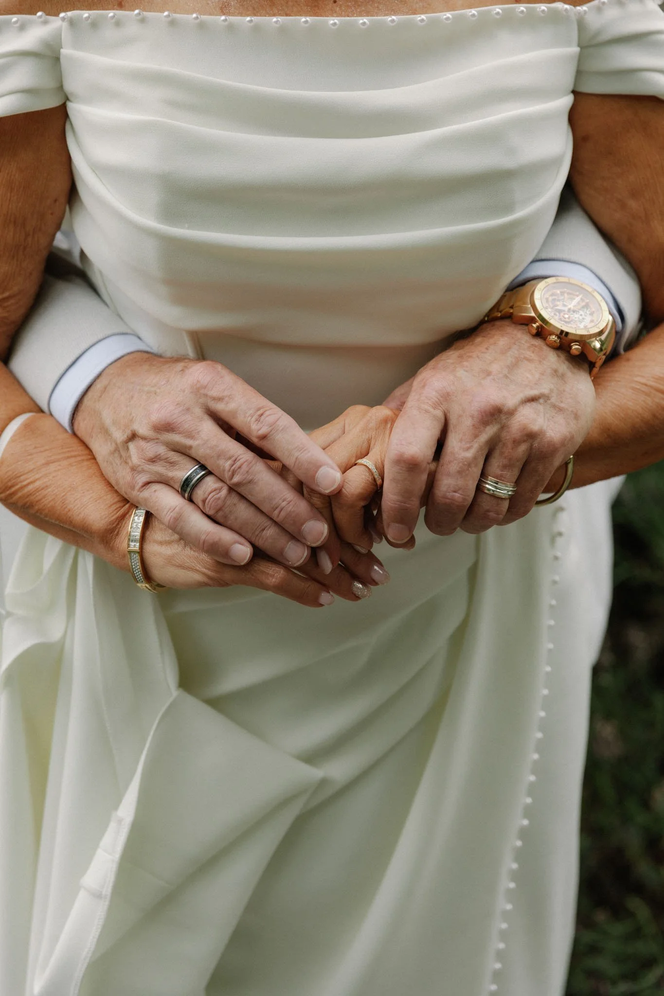 A close-up of a person's hands, adorned with multiple rings, holding another person's hand. The person being held is wearing a bright watch with a large face. Both individuals are dressed in elegant attire, with the person in the background wearing a white dress with structured, pleated fabric and pearl embellishments along the neckline, likely at a formal event such as a wedding.