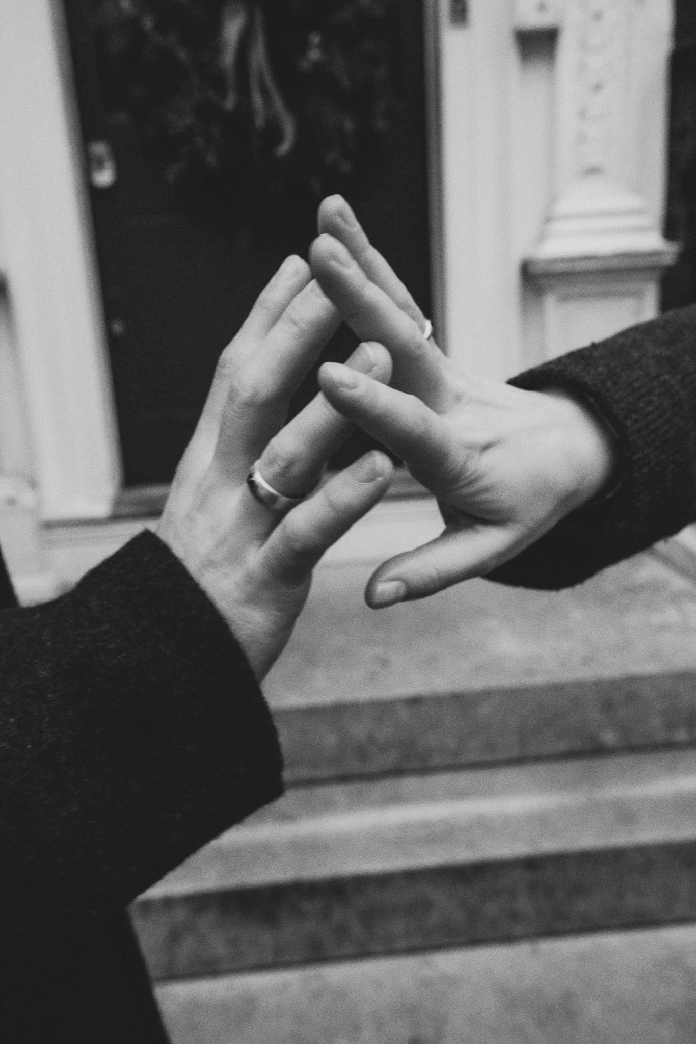Black and white photo of two hands touching, with fingers interlaced, near a mirror. One hand has a ring on the ring finger. The background shows a window and part of a house interior.