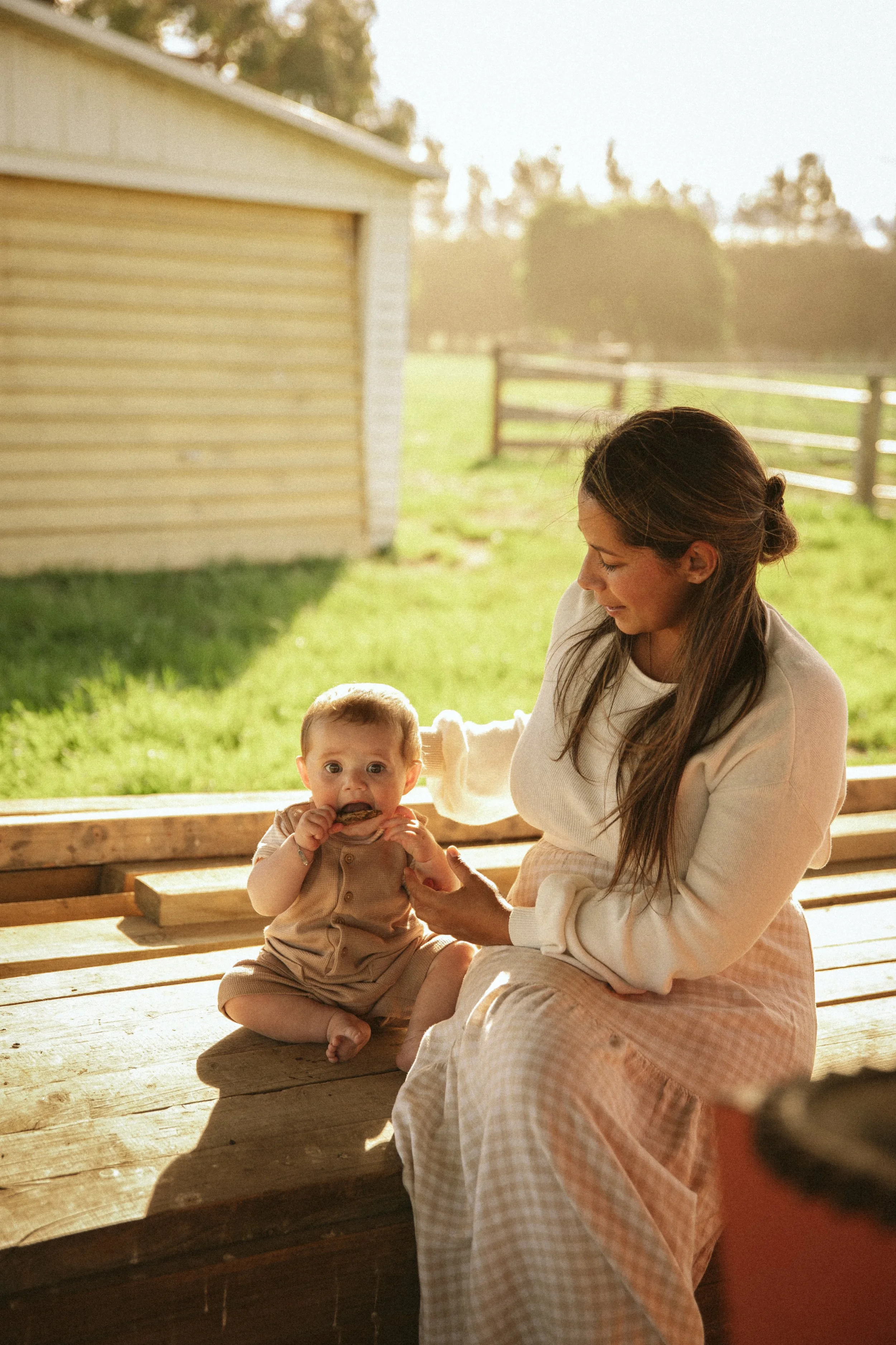 Woman and baby sitting outdoors on a wooden surface, near a barn, with a grassy field and wooden fence in the background. The woman is wearing a light sweater and skirt, while the baby is dressed in a beige outfit, holding a leaf.