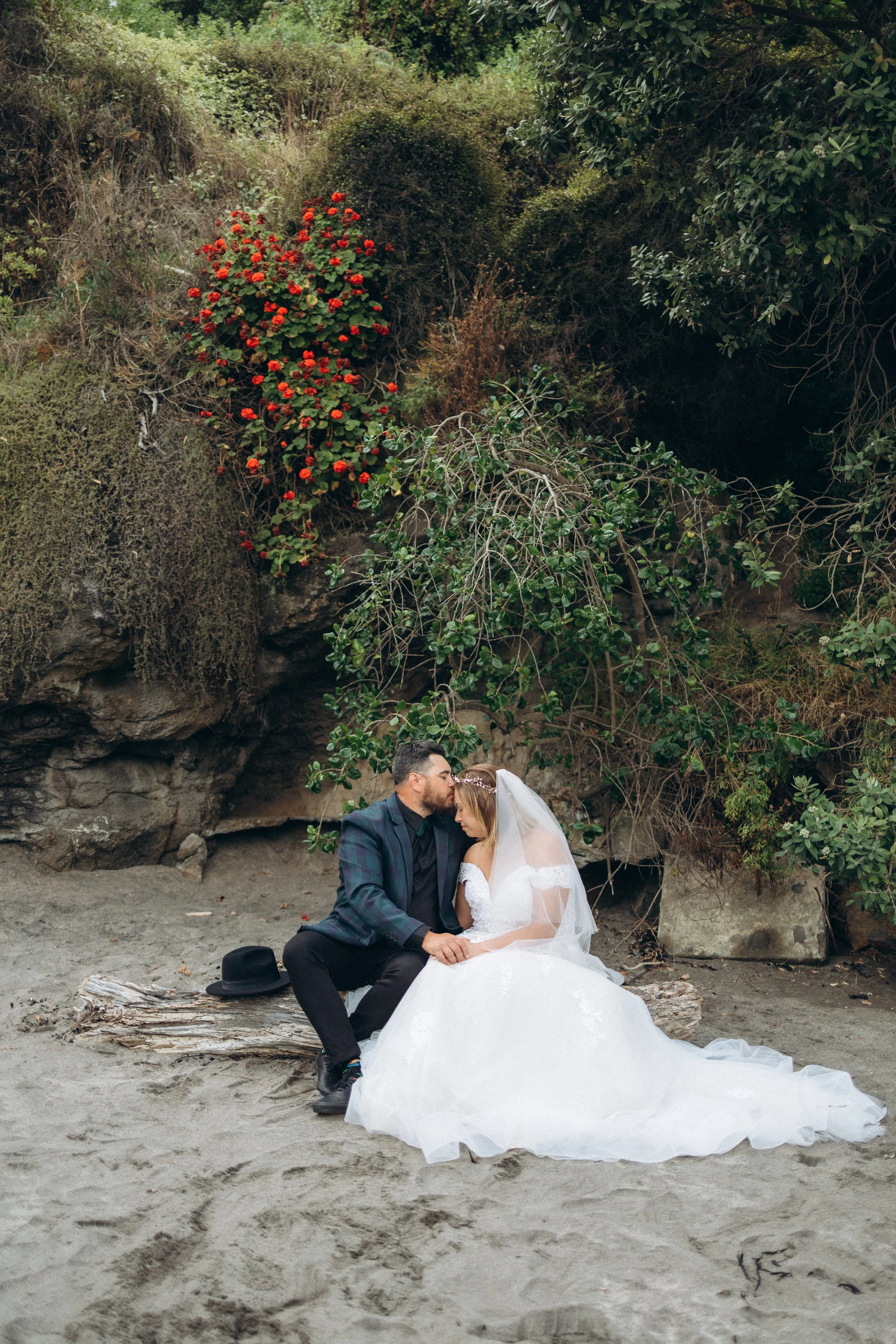 Bride and groom sitting on a beach surrounded by greenery and red flowers, with the groom kissing the bride's forehead.
