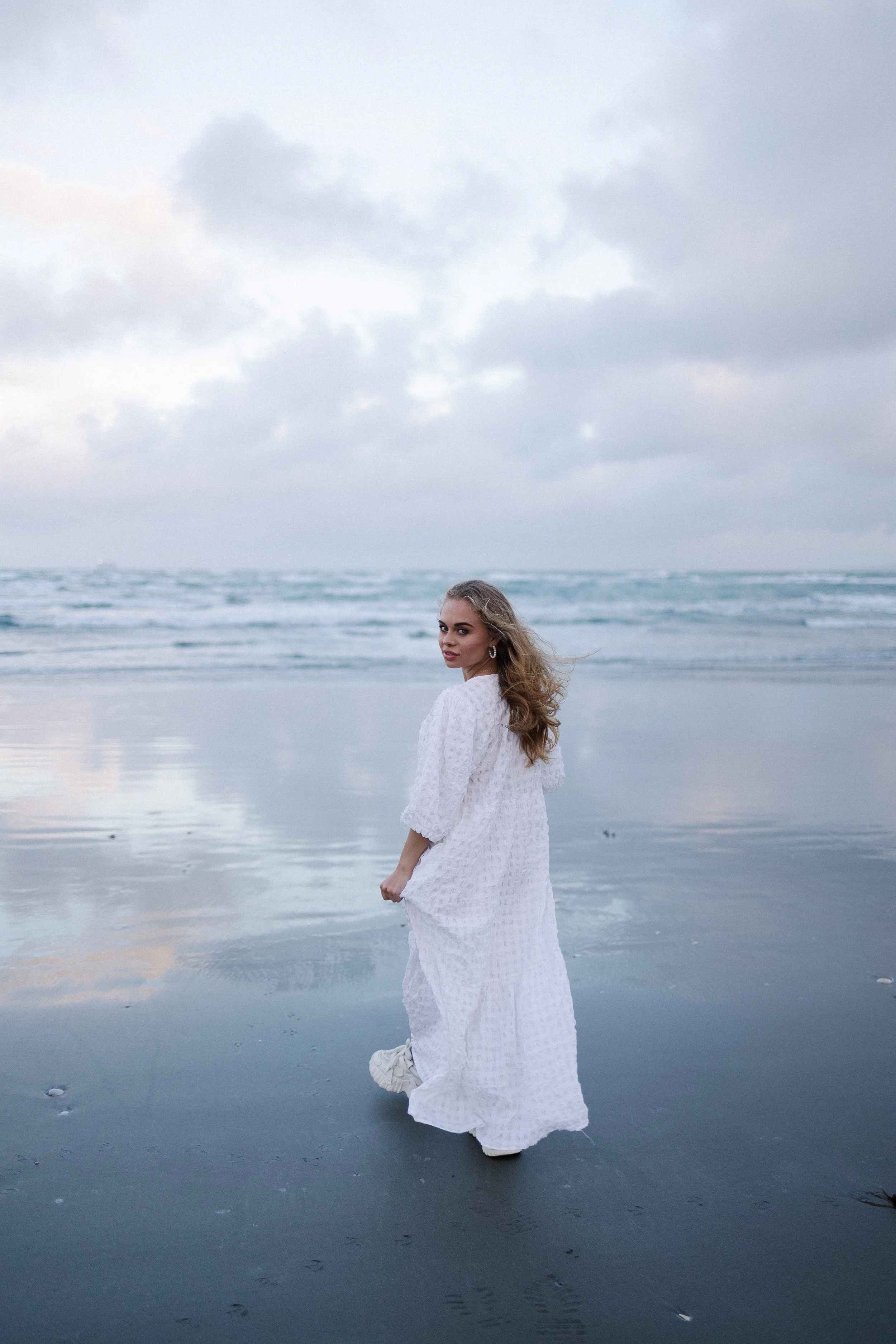 Woman in a white dress walking on a beach at sunset with ocean waves in the background.