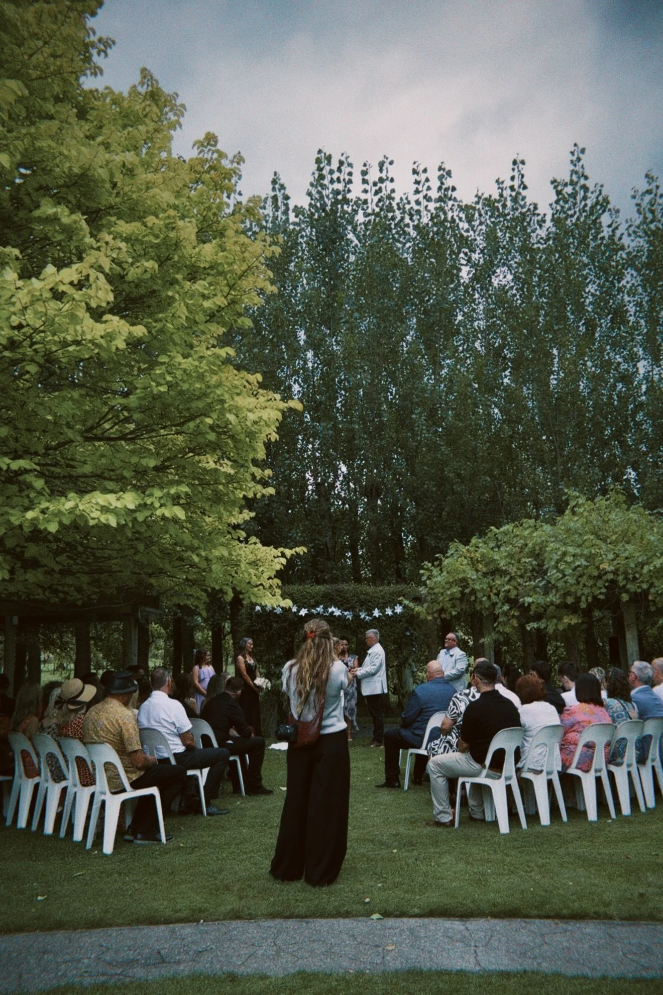 An outdoor wedding ceremony with guests seated on white chairs under a canopy of green trees in a garden setting. A small group of people, including the bride and groom, are standing near the altar, with the officiant. The scene appears to be in the evening.