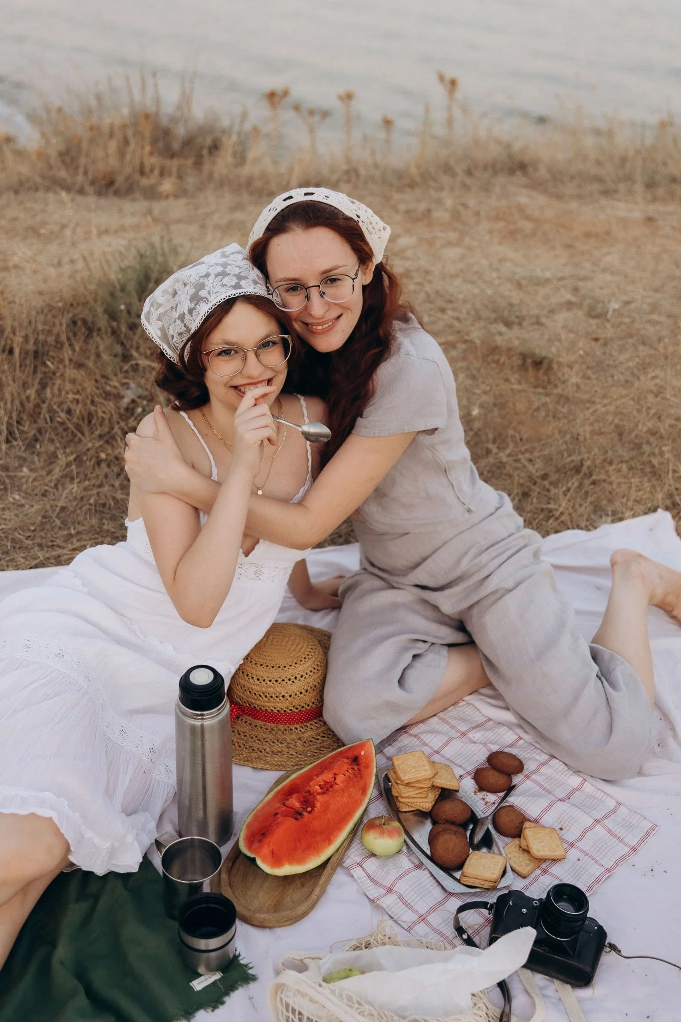 Two women with glasses, wearing white lace head coverings and light-colored dresses, smiling and hugging while sitting on a blanket during a picnic near a body of water. There are picnic items around them, including a watermelon, sandwiches, cookies, apples, thermoses, a camera, and a straw hat.