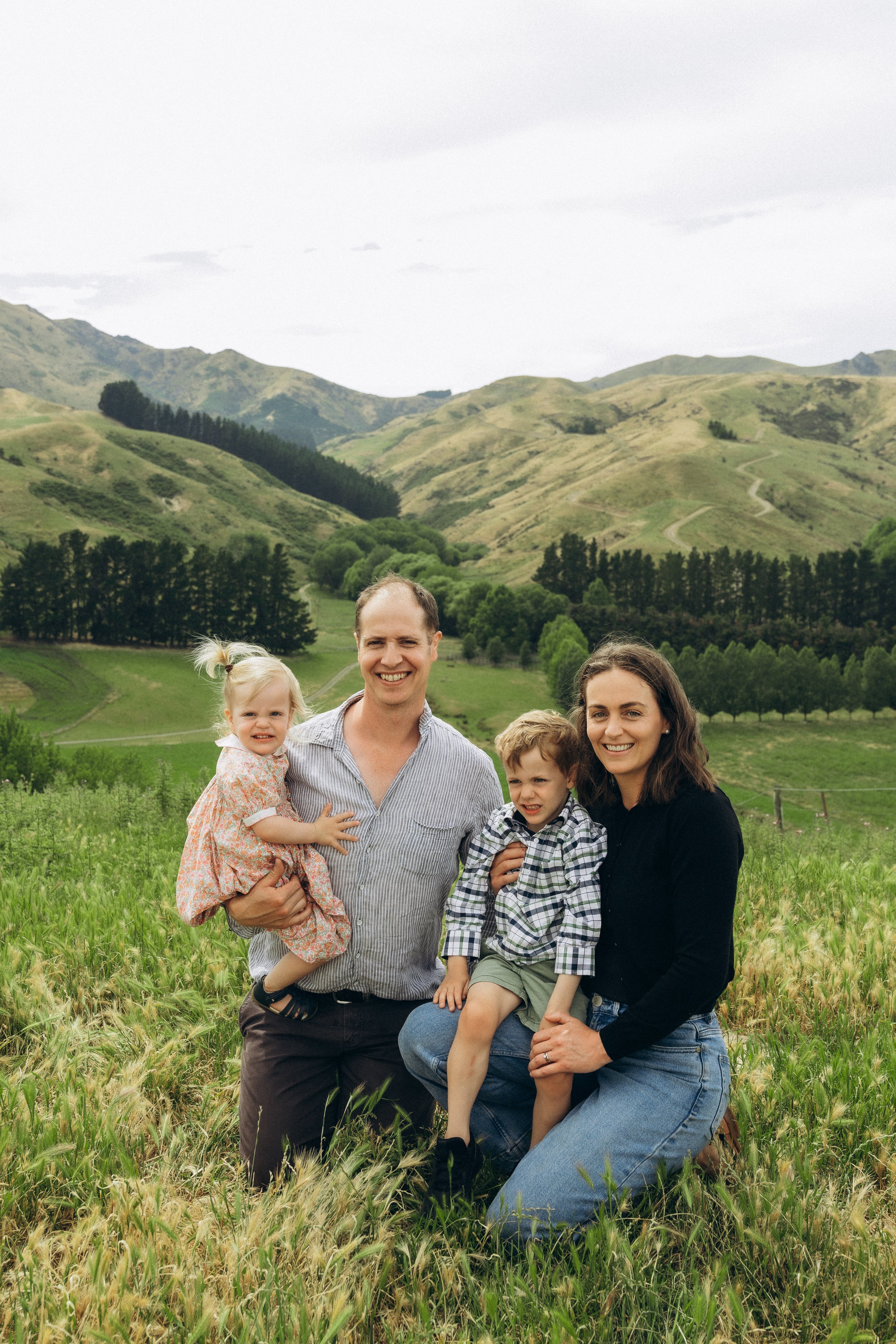 A family of four poses in a grassy field with rolling hills and trees in the background. The parents, a man and a woman, kneel in the grass while holding a young girl and boy.