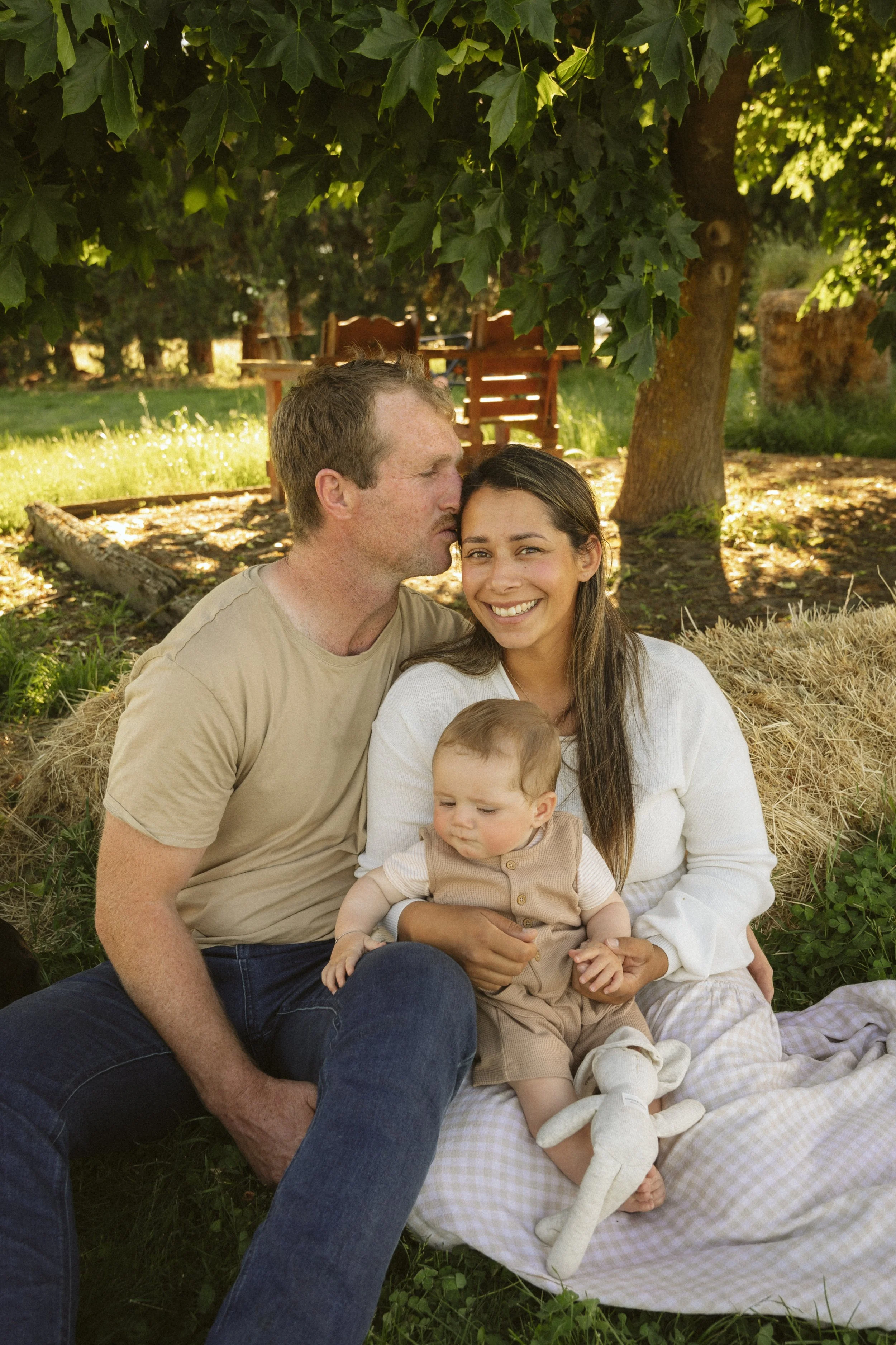A family of three, including a baby, sitting on a picnic blanket in a park. The man kisses the woman on the forehead, who smiles at the camera while holding the baby. They are sitting under trees with hay bales nearby.