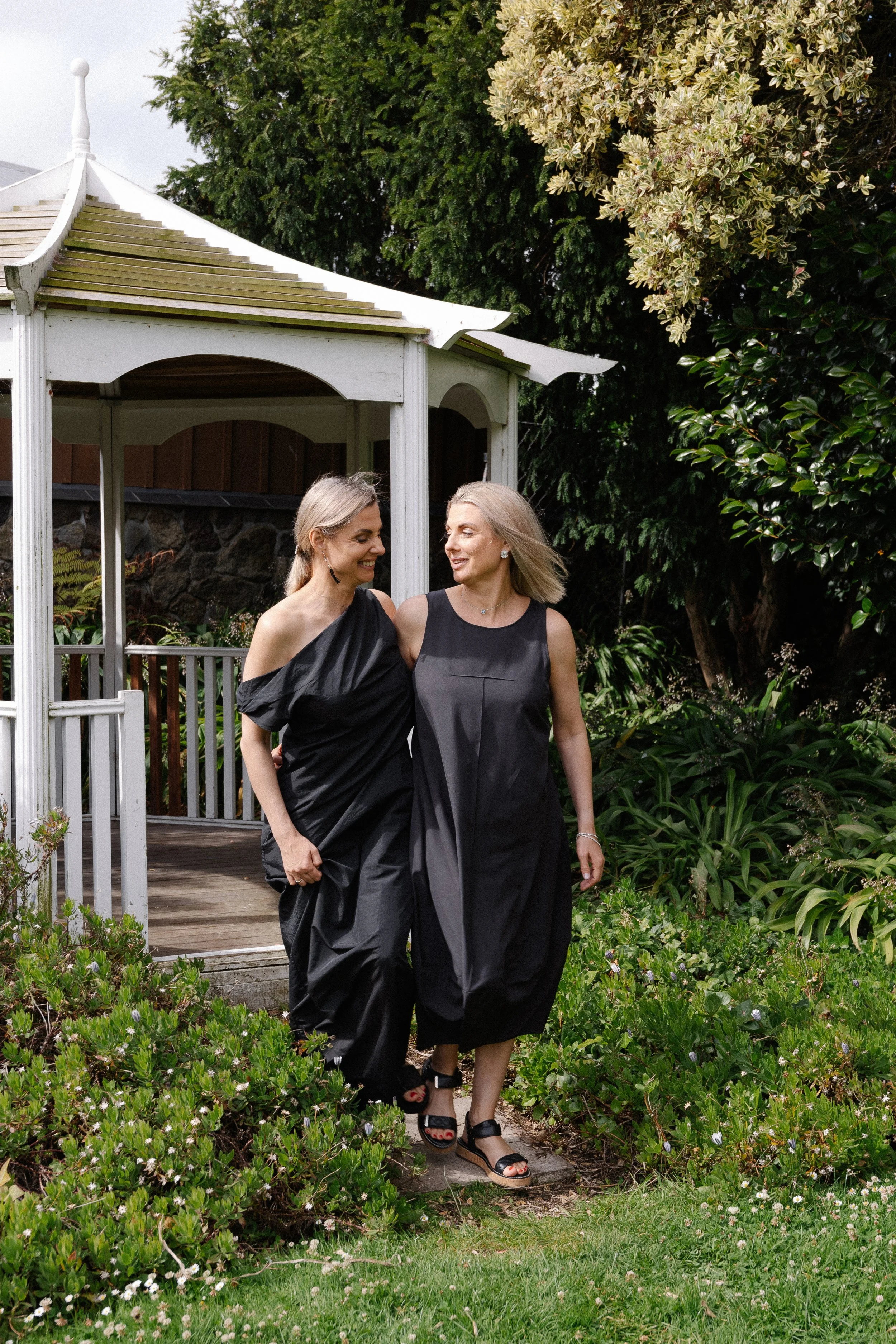 Two women in black dresses walking in a garden, near a white gazebo, surrounded by greenery.