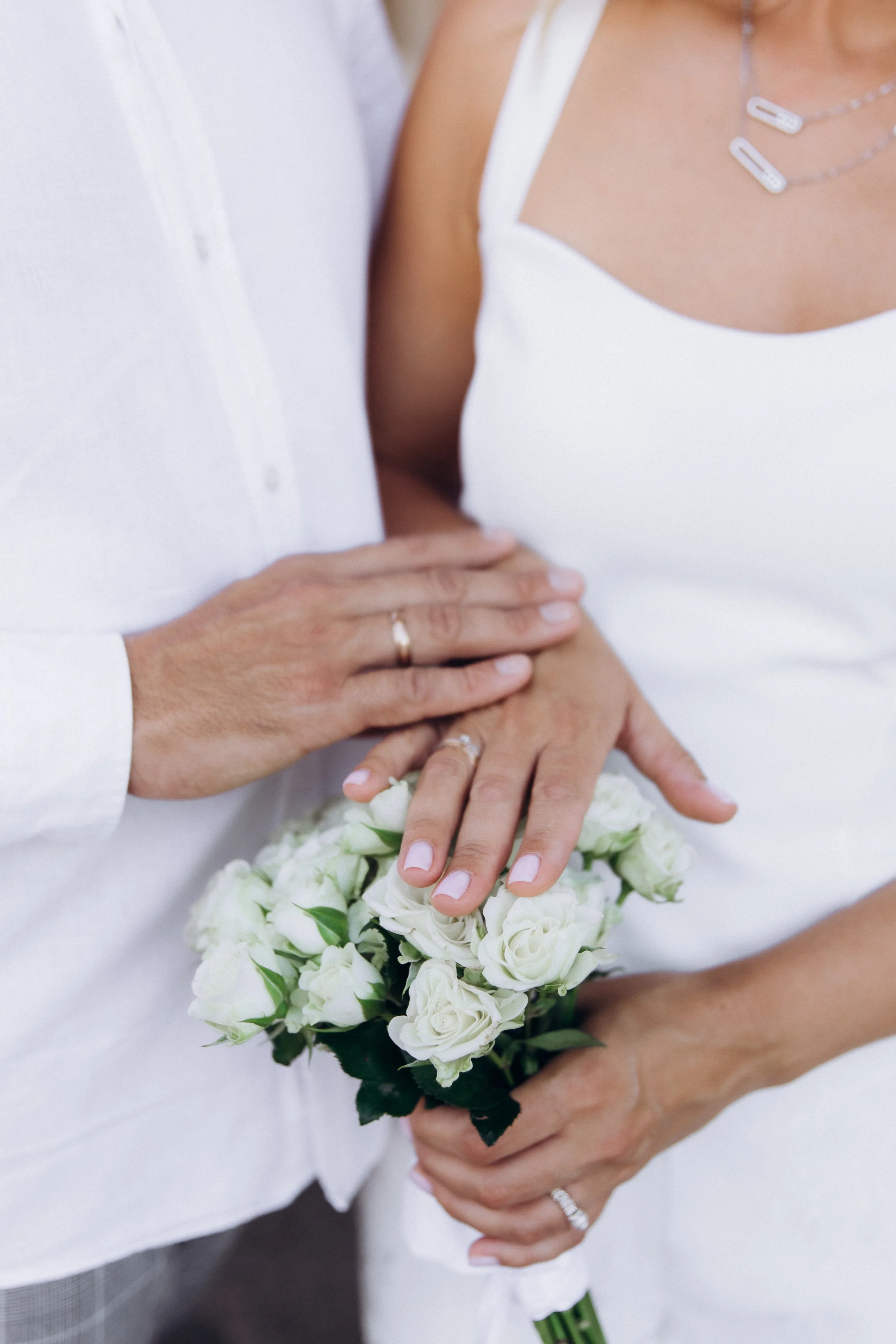 Close-up of a bride and groom's hands, with the bride holding a bouquet of white roses, and both wearing wedding rings, during their wedding celebration.