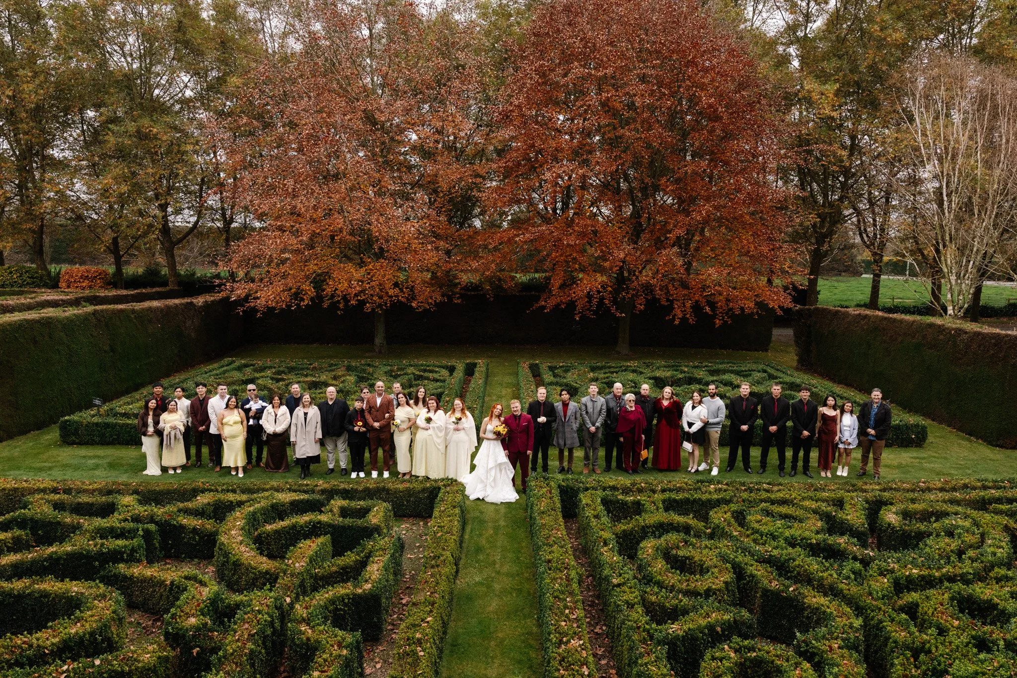 Group of people gathered outdoors in a garden with manicured hedges and trees with fall foliage.