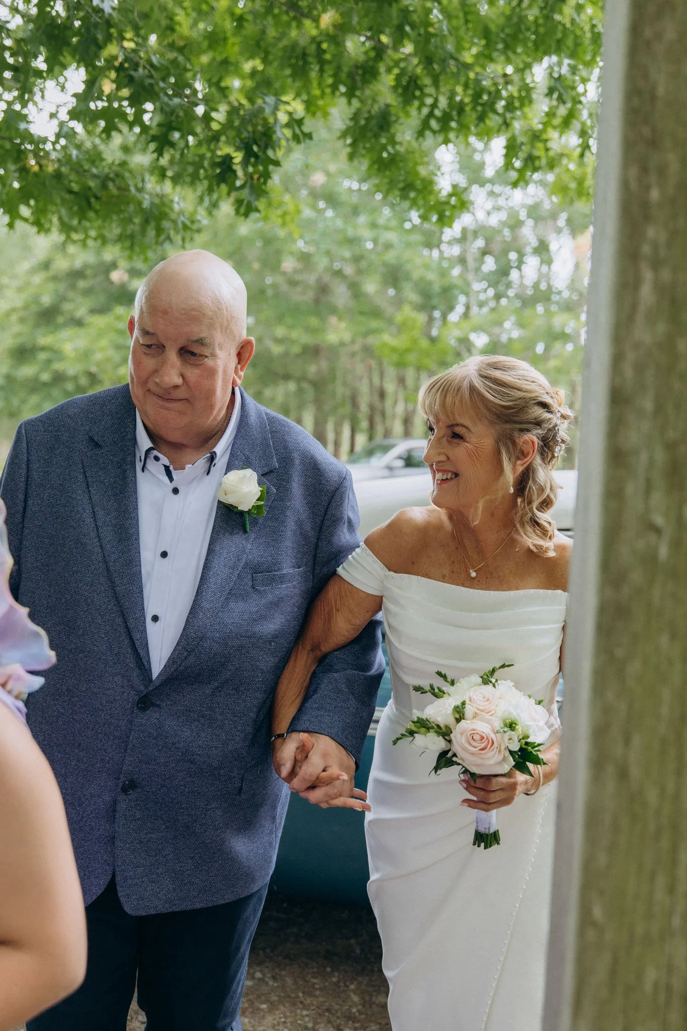 A bride holding a bouquet of pink and white roses, smiling, standing next to a groom with a bald head, wearing a gray suit and white shirt, during a wedding ceremony outdoors under green trees.