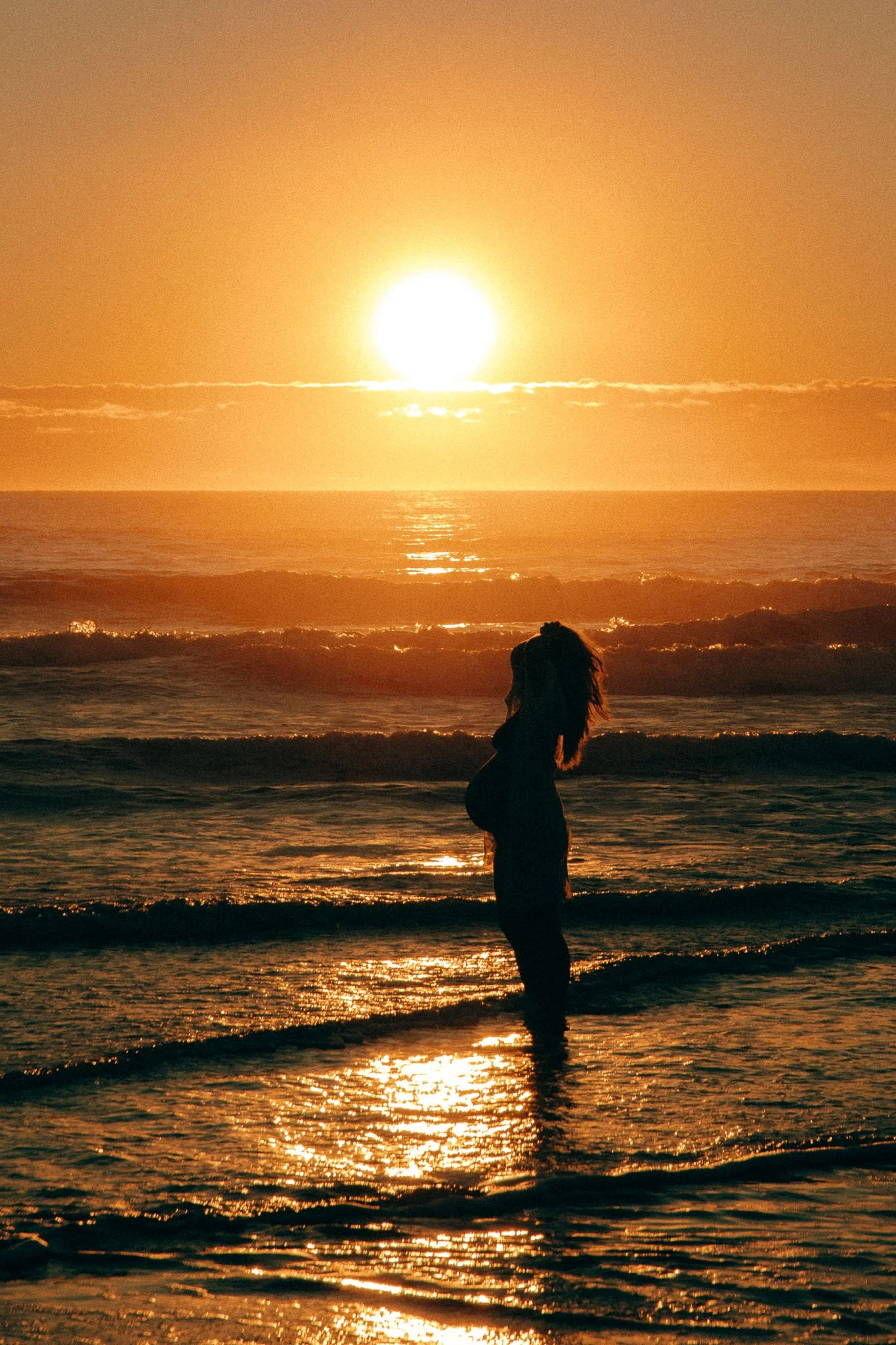 A person standing on the beach during sunset, with the sun low on the horizon reflecting off the water and the person's silhouette visible.