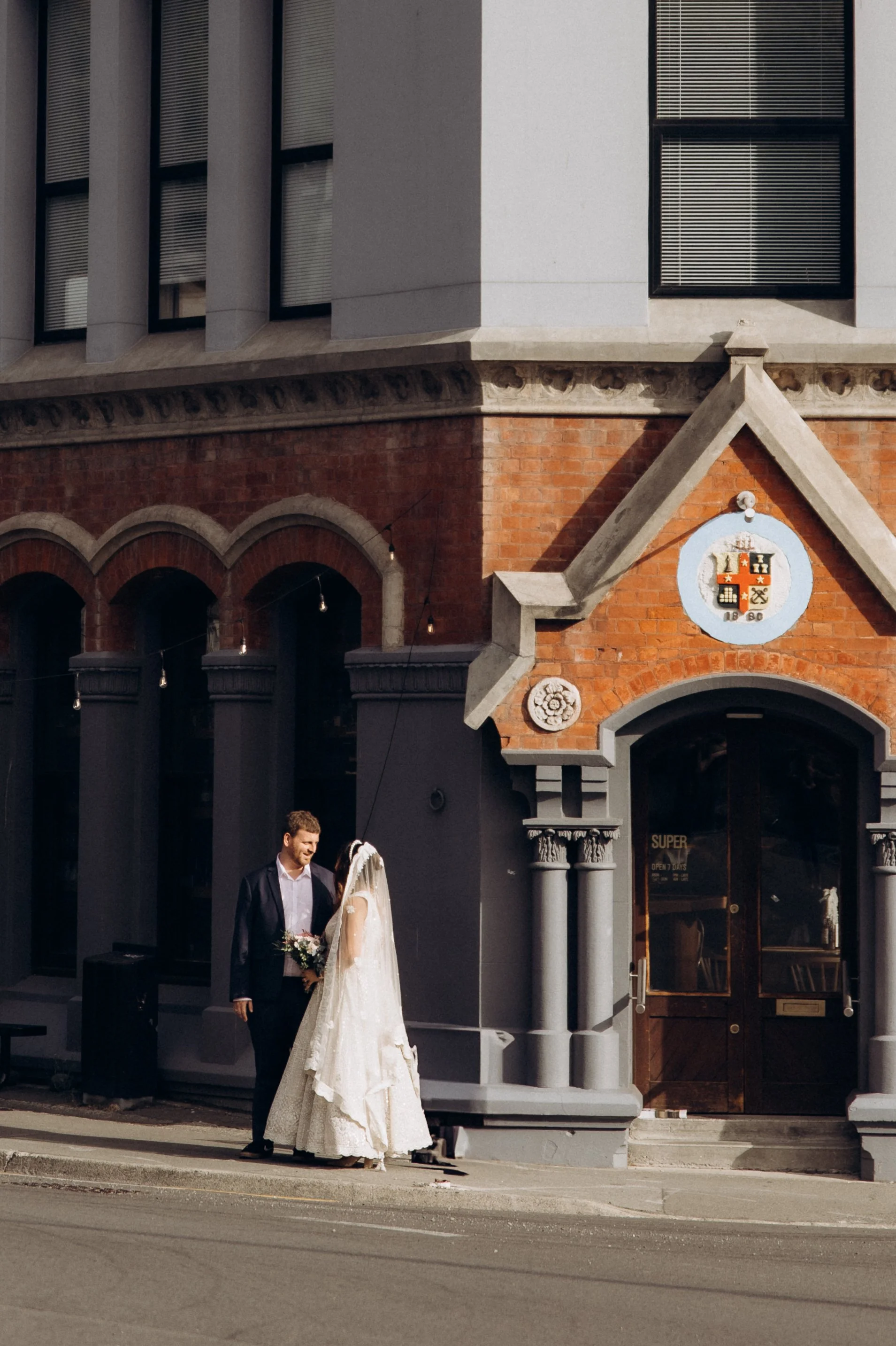A bride and groom standing on a sidewalk outside a brick building, with arched windows and a crest above the entrance. The groom is in a suit, and the bride is in a white wedding dress, holding a bouquet.