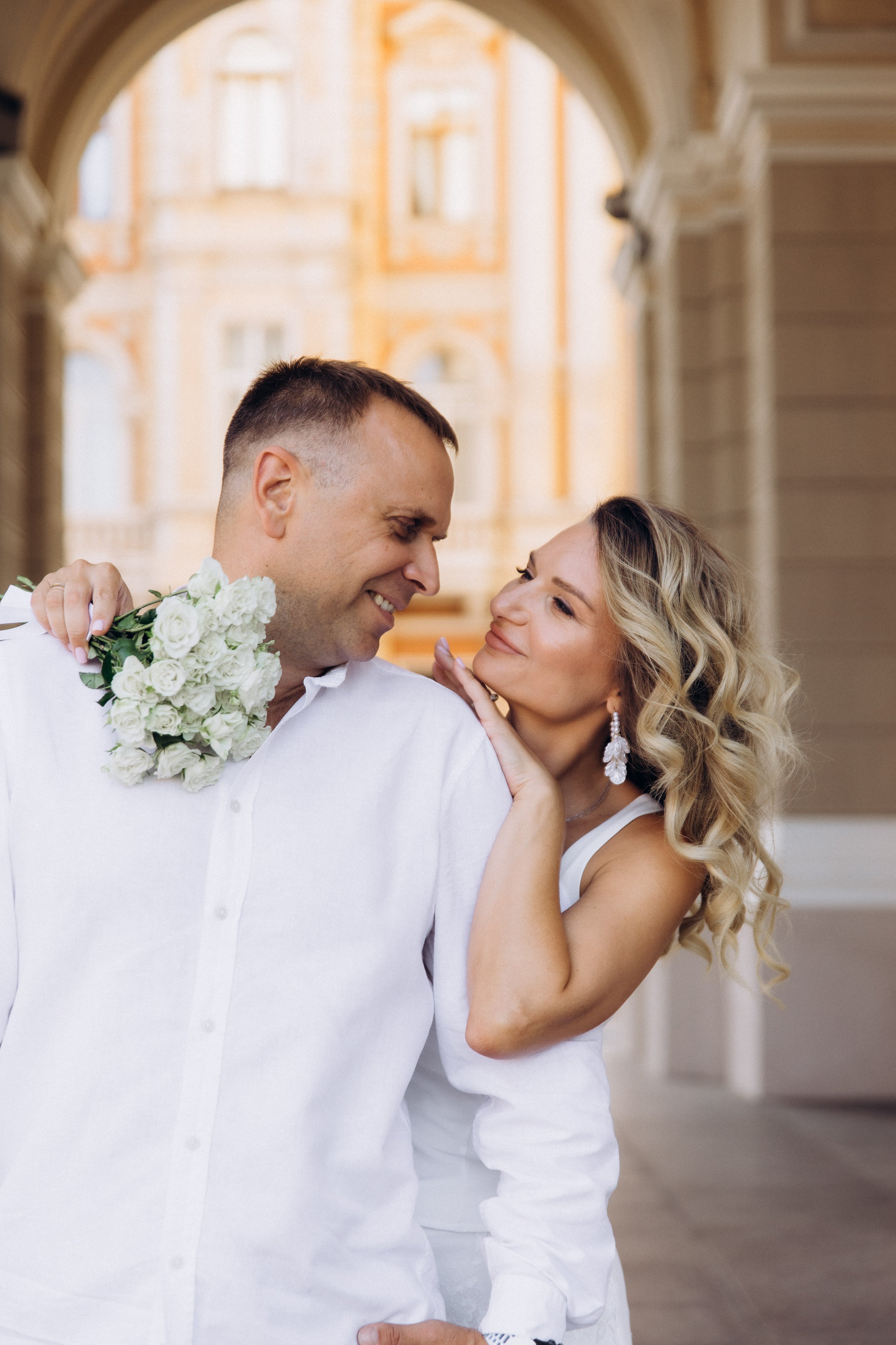 A couple on their wedding day smiling at each other, with the woman touching the man's face and the man holding a bouquet of white flowers, under an archway with a large building in the background.