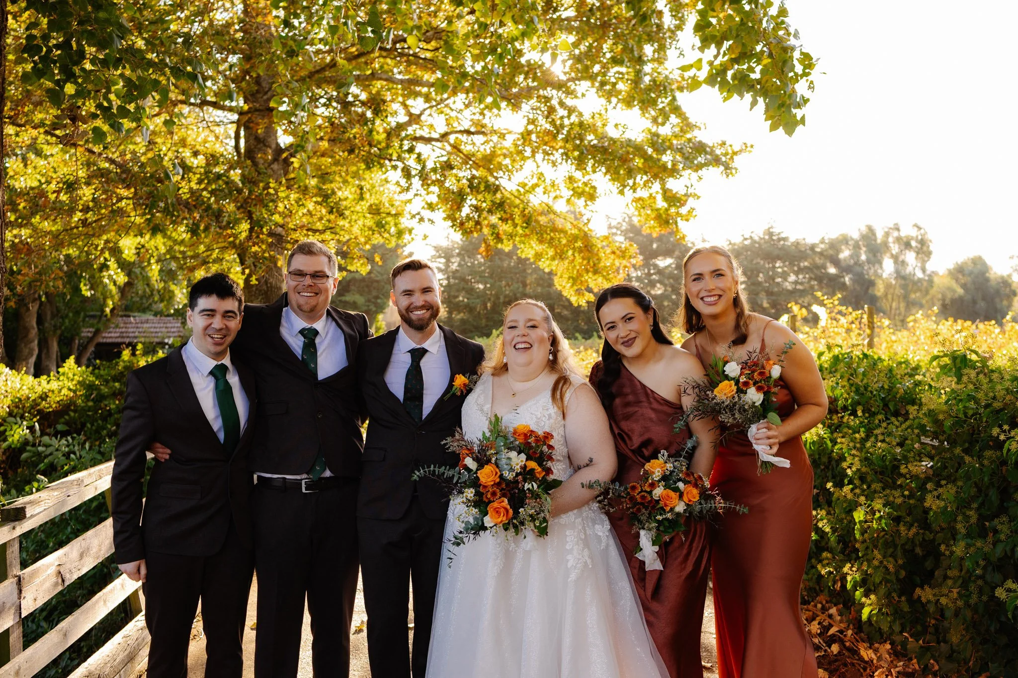 A wedding group photo of six people standing outdoors, surrounded by trees with sunlight filtering through the leaves, holding bouquets with orange, yellow, and white flowers, on a wooden pathway.
