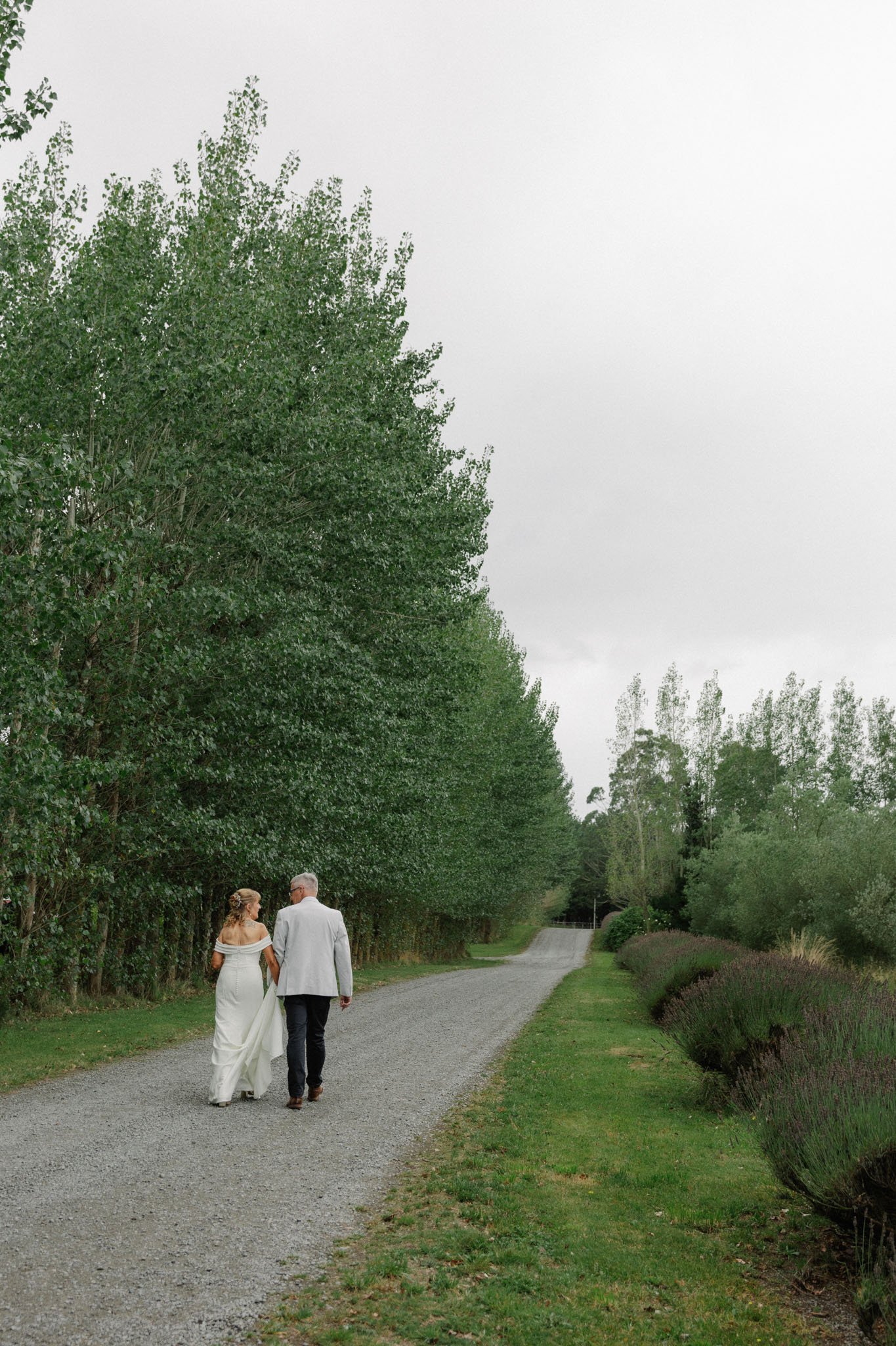 A bride and groom walking hand in hand down a gravel path lined with green trees and bushes on a cloudy day.