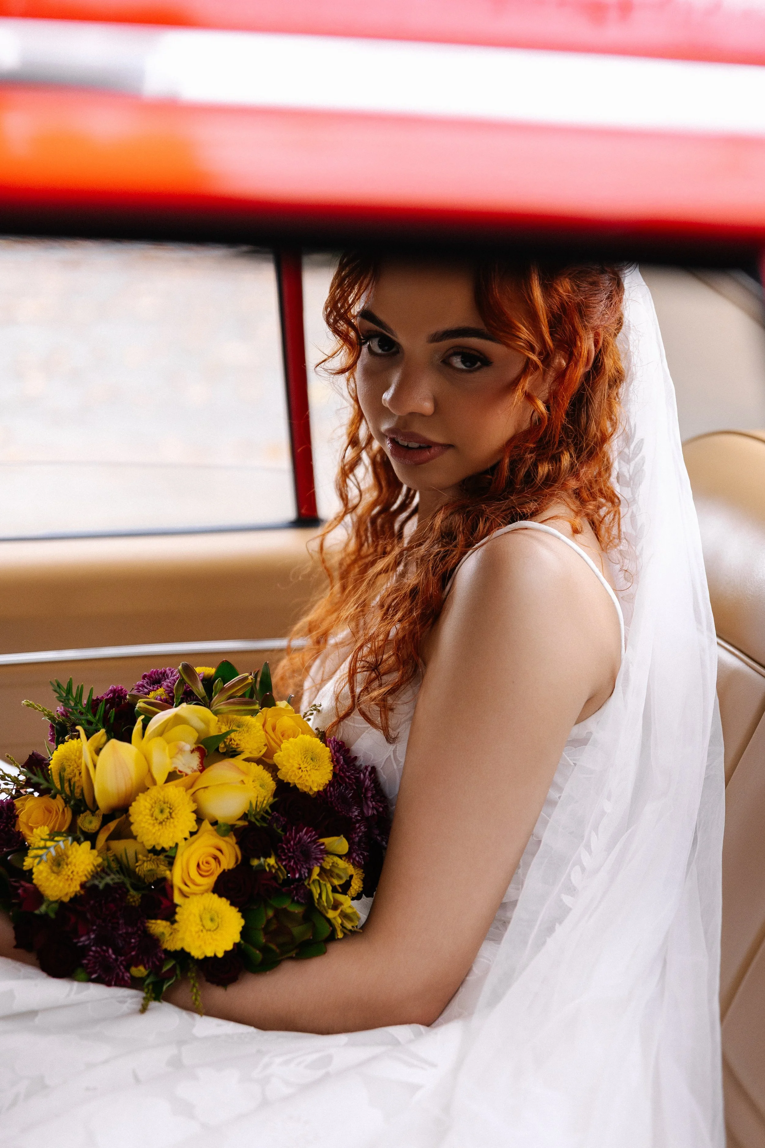 A woman with red curly hair wearing a white dress, sitting on a vintage car, holding a bouquet of yellow and purple flowers.