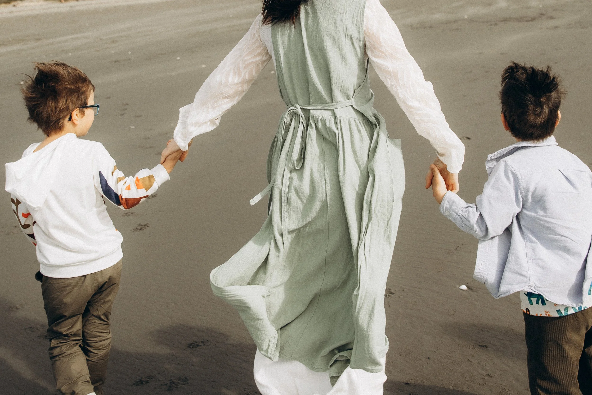 Back view of a woman holding hands with two children on a sandy beach.