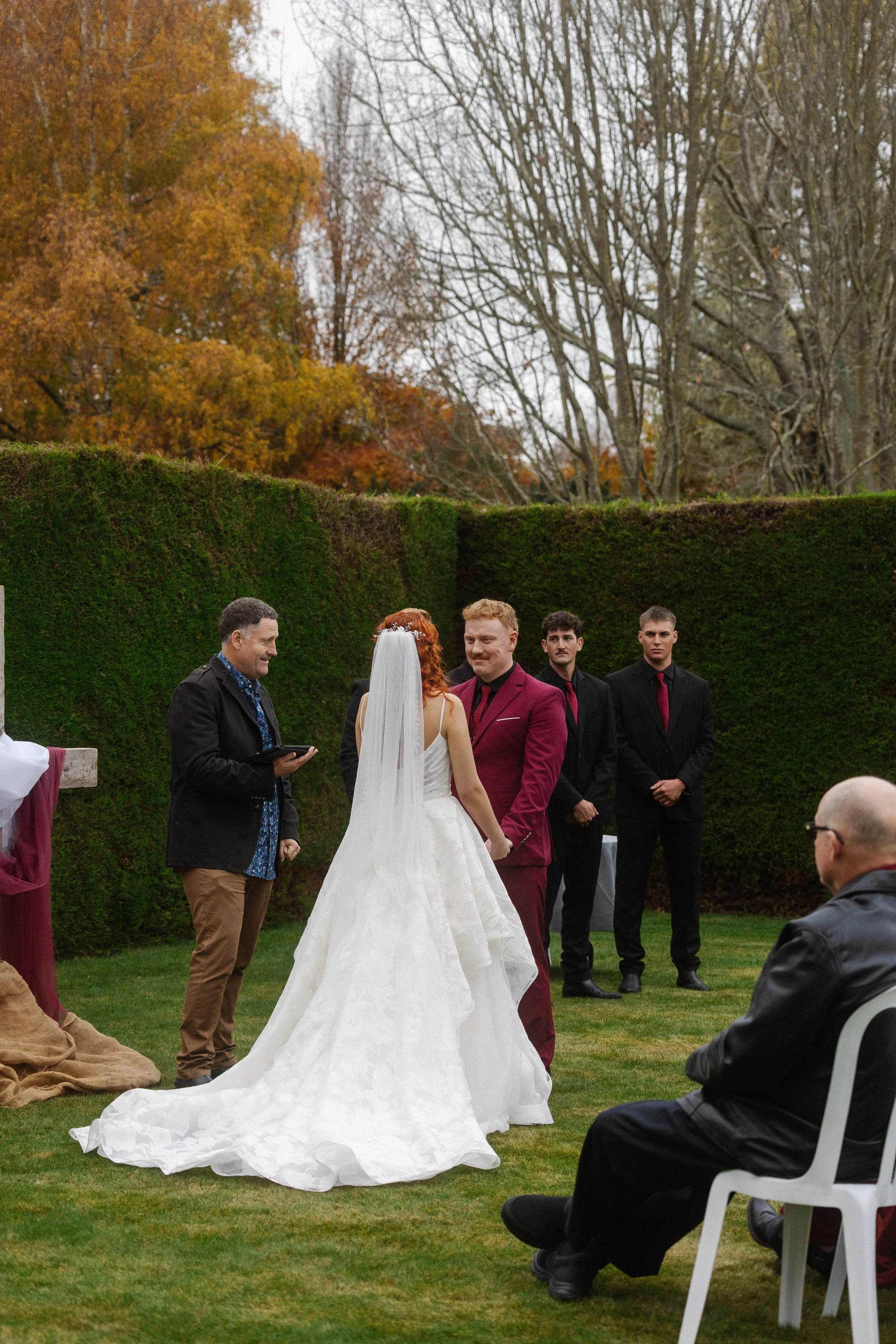 A wedding ceremony outdoors with a bride in a white gown and veil, a groom in a burgundy suit, and officiants and guests, set against a backdrop of green hedge and autumn trees.