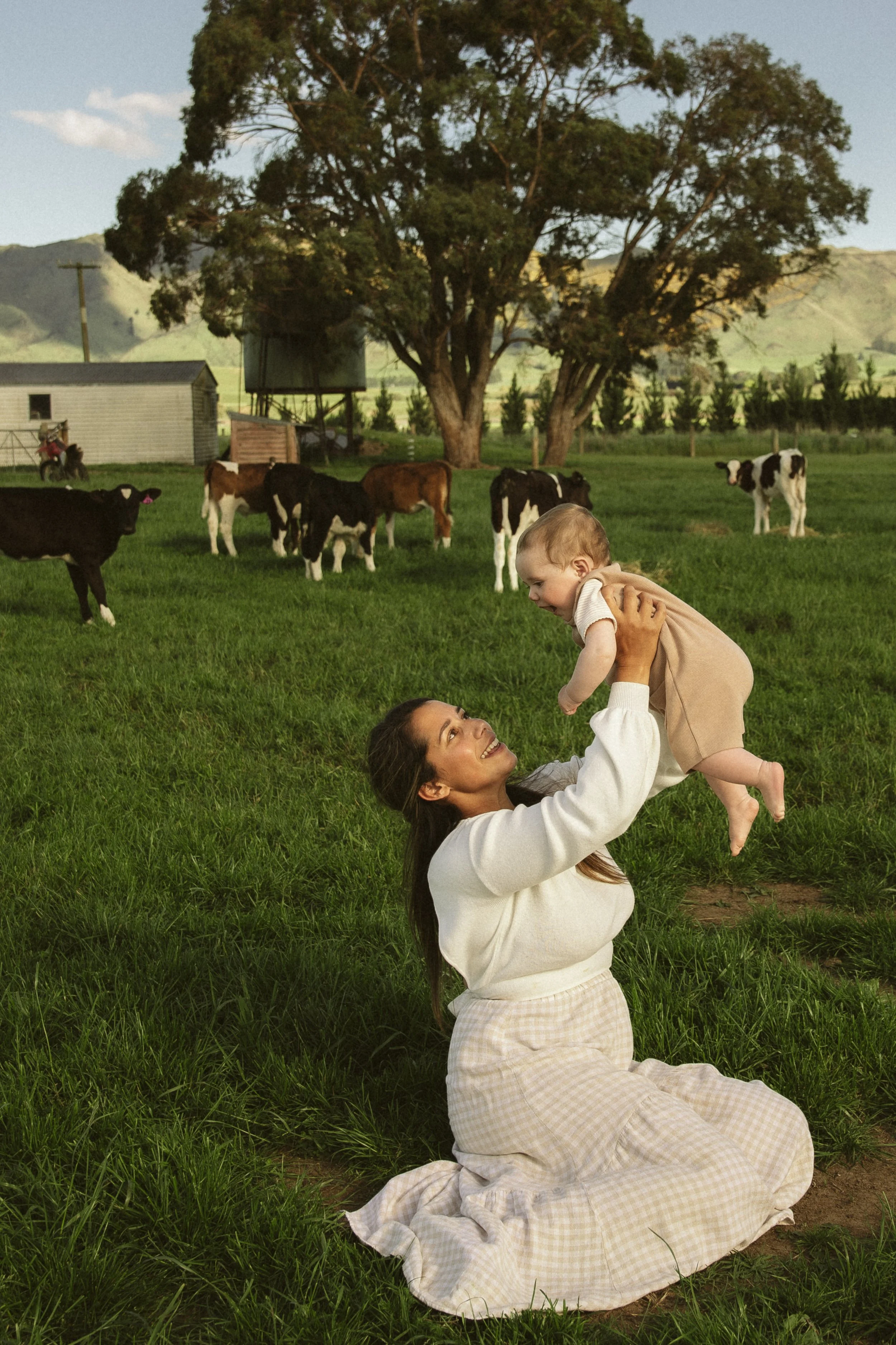 Woman holding a baby in a grassy field with cows, trees, and mountains in the background.