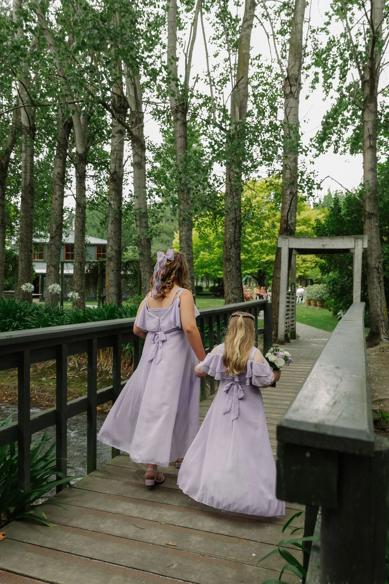 A woman and a young girl, both wearing matching lavender dresses, walk hand in hand across a wooden bridge in a lush, green park. The woman has a large lavender bow in her hair and carries a bouquet of flowers, suggesting they are participating in a wedding.