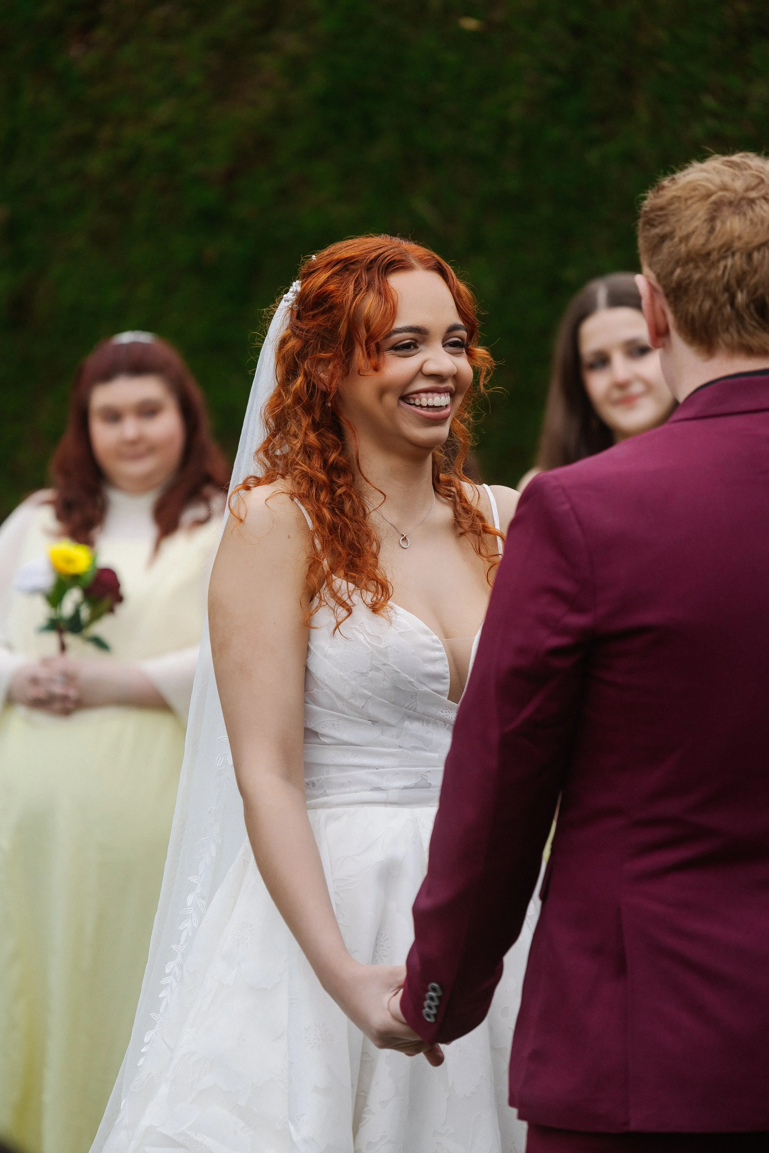 A bride and groom holding hands during a wedding ceremony outdoors. The bride, with red curly hair, is smiling at the groom, who has light hair. In the background, three women are visible, one holding a bouquet of yellow and dark red flowers, and the