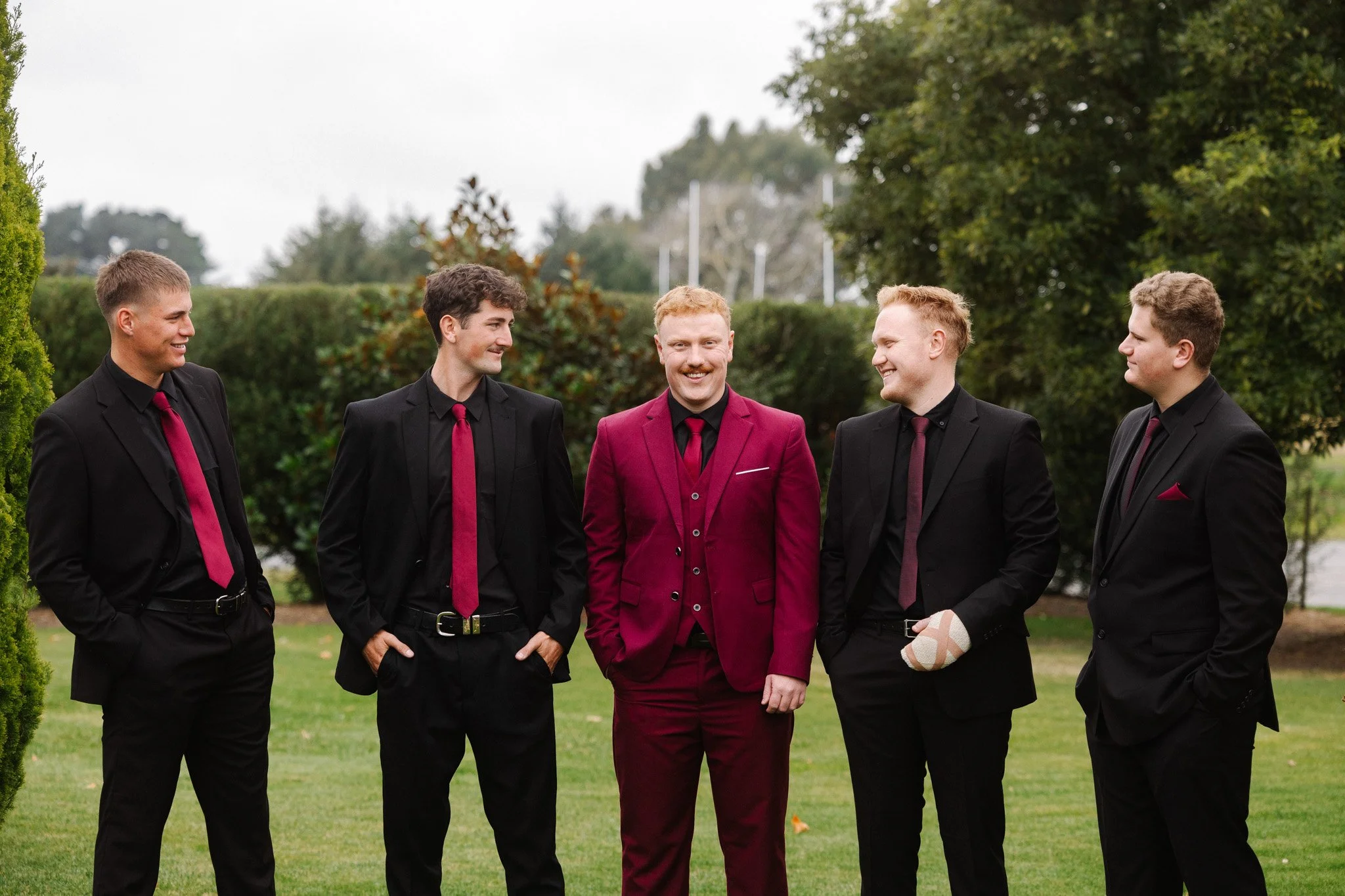 Group of five men dressed in black suits with burgundy ties, standing outdoors on a grassy area with trees in the background, smiling and interacting with each other.