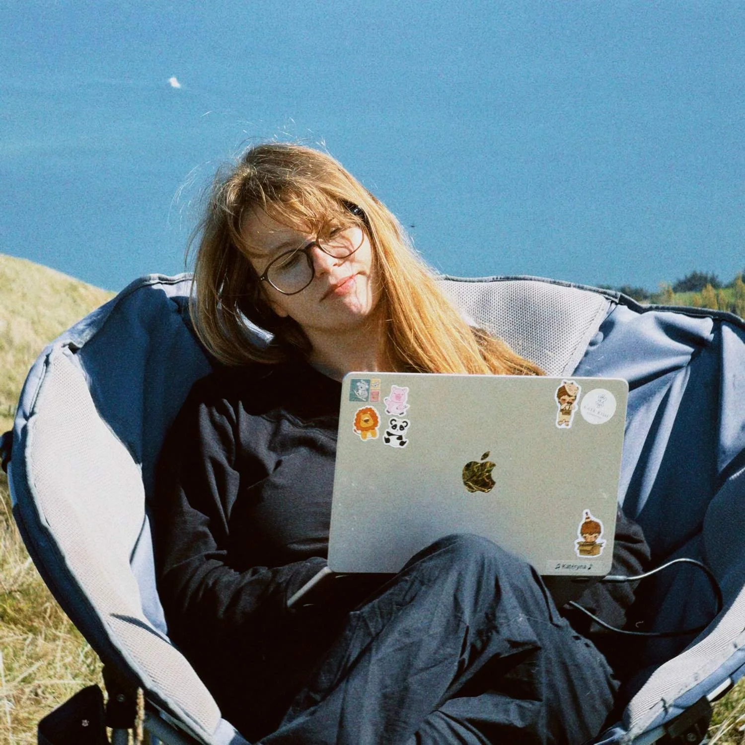 A young woman with red hair, glasses, and a black jacket sitting outdoors on a camping chair, looking at a laptop decorated with colorful stickers, with a blue sky and green landscape in the background.