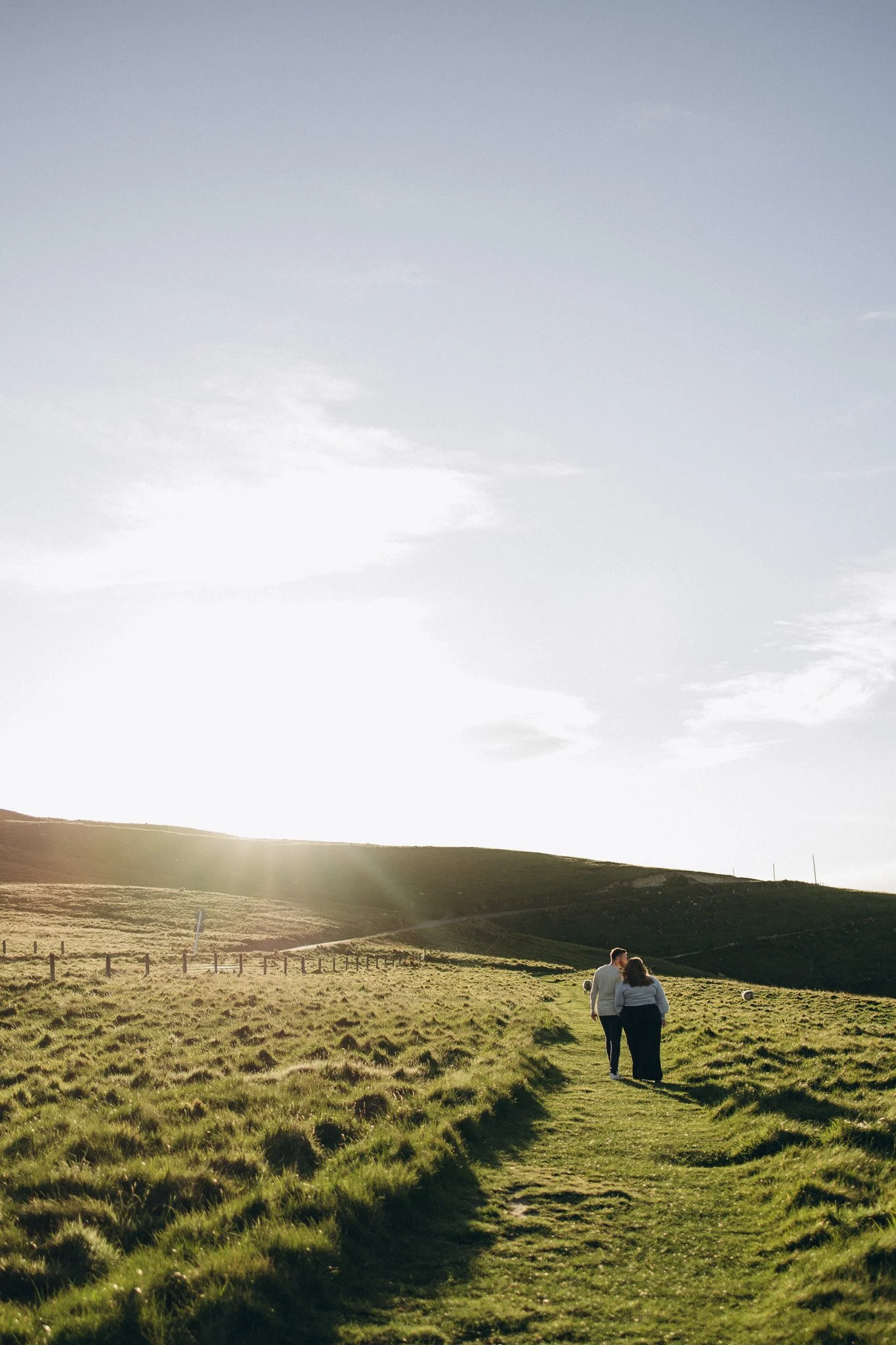 A couple walking along a grassy trail in a countryside landscape during sunset, with a fence and hills in the background, and a few sheep grazing nearby.
