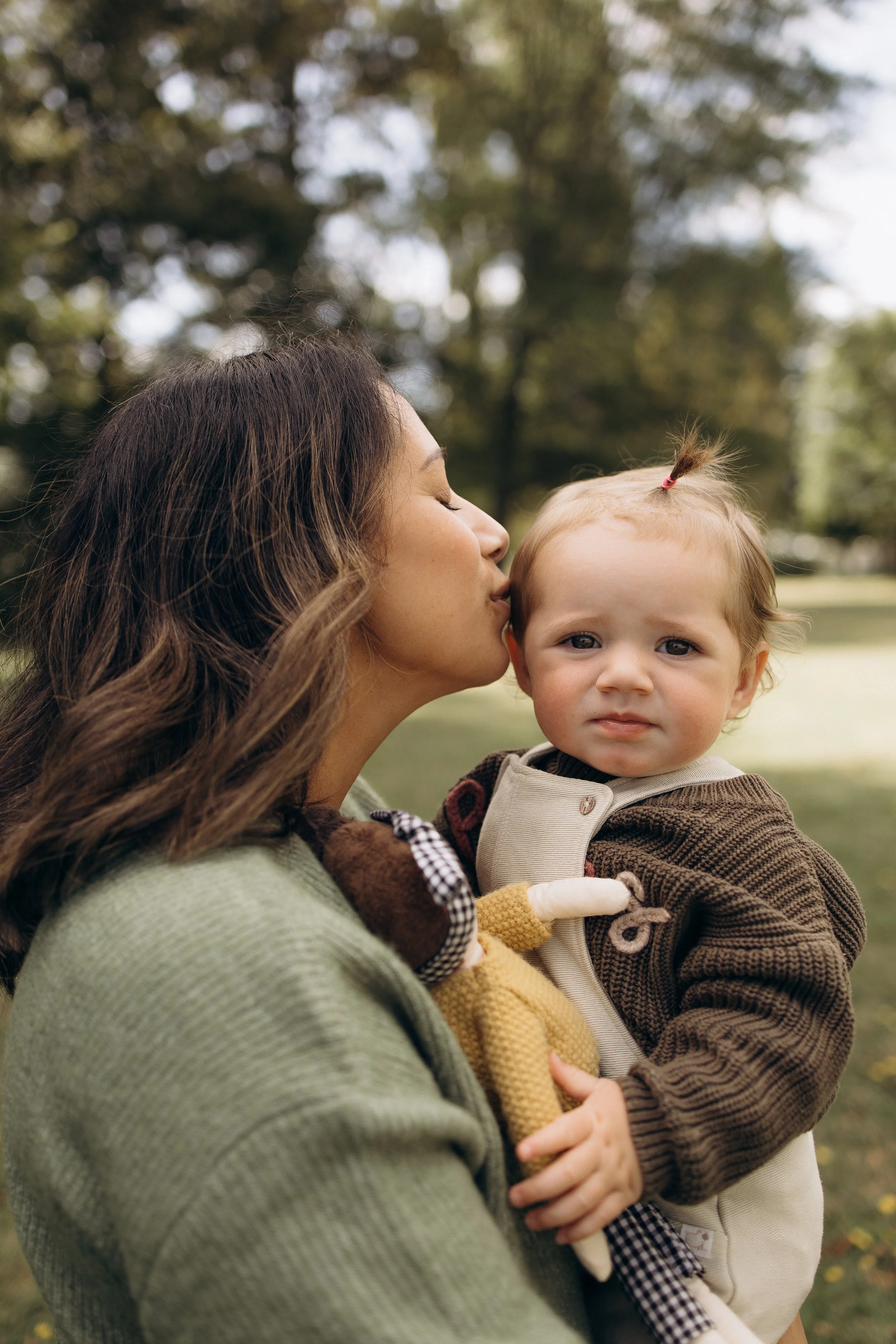 A woman kissing a child on the cheek while holding a doll, in an outdoor setting with trees in the background.