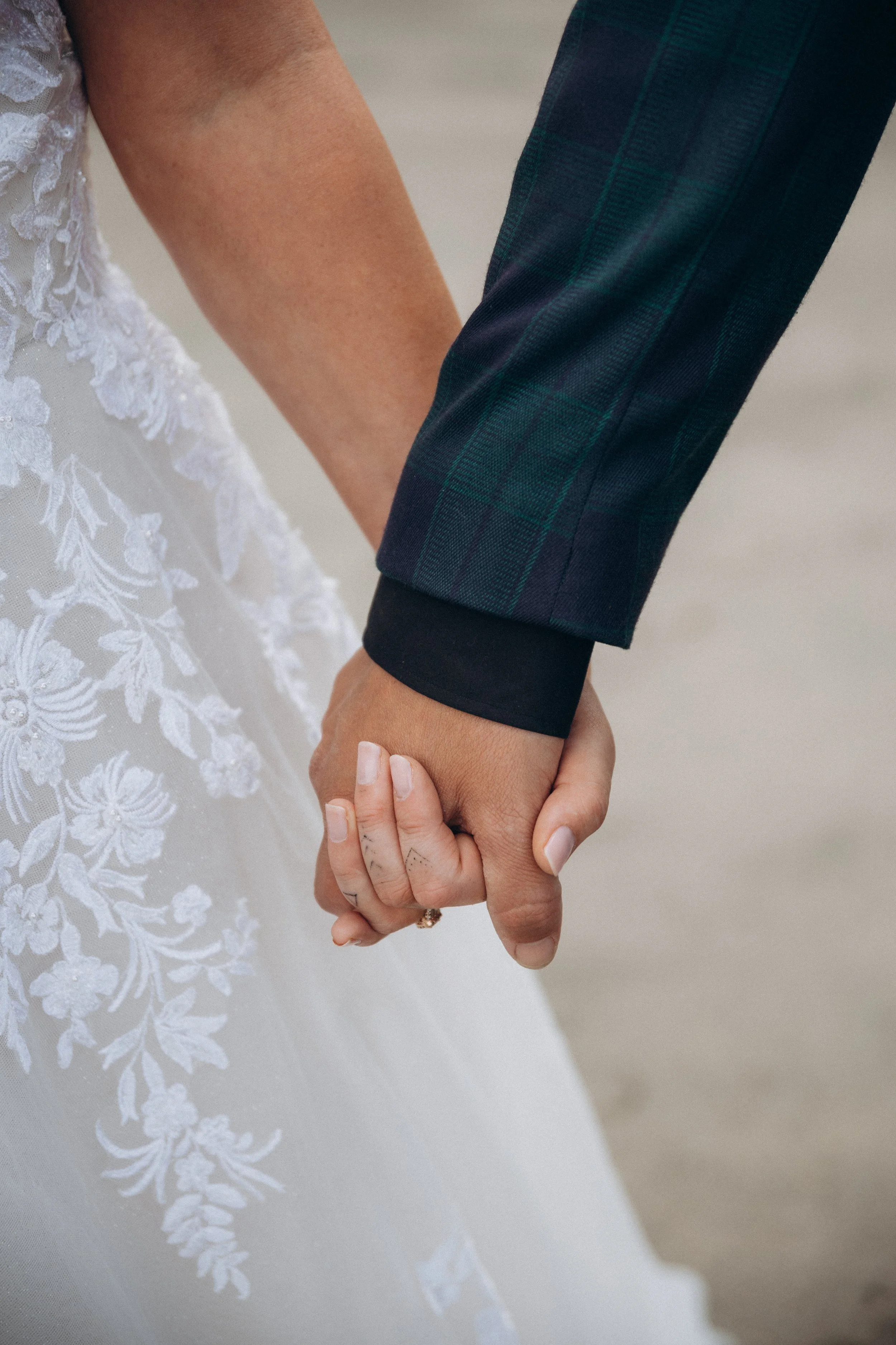 Close-up of a couple holding hands, with one wearing a wedding dress featuring floral lace and the other in a dark suit sleeve.