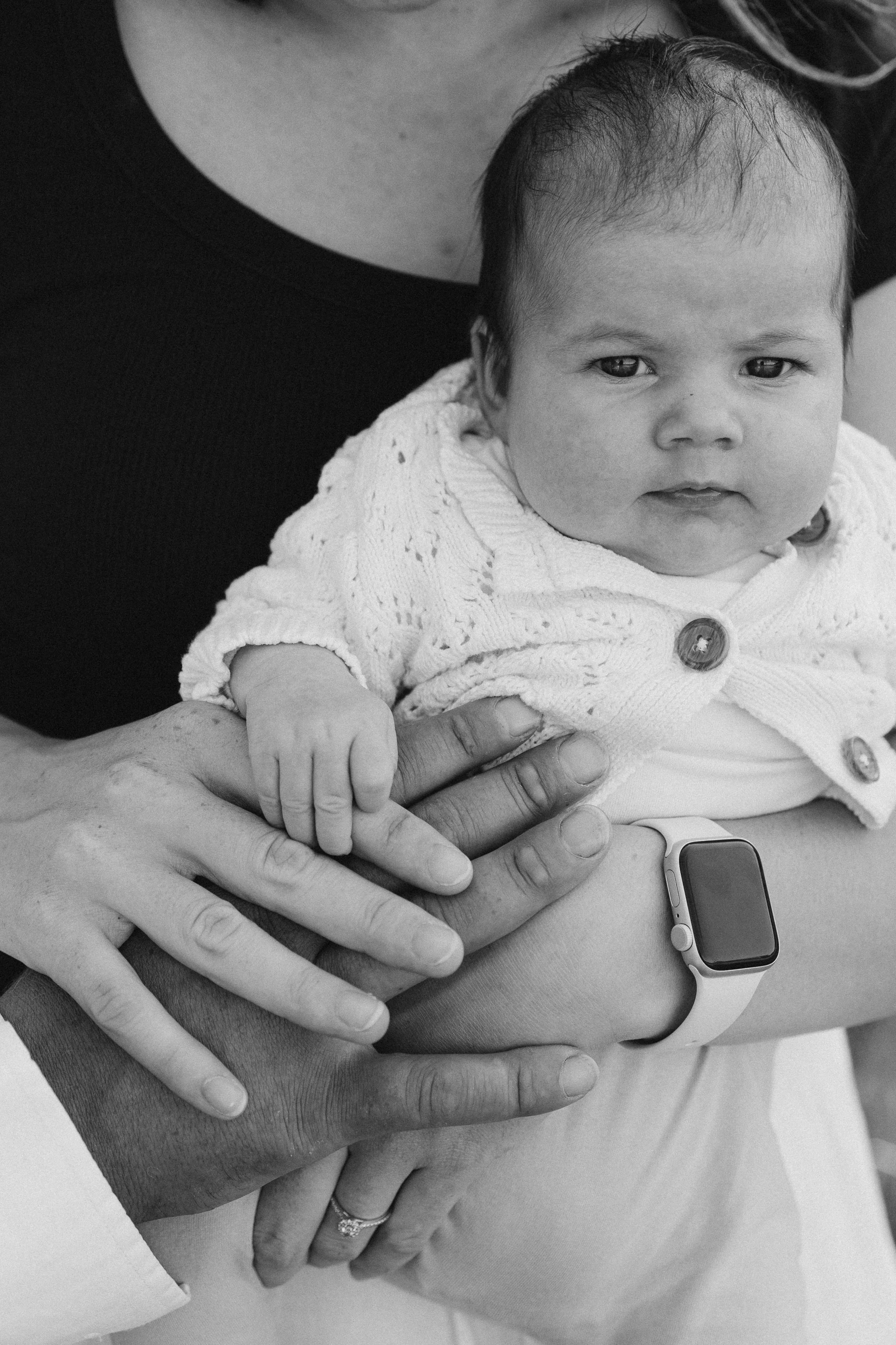 A baby in a white knitted sweater being held by two adults. One adult is wearing a smartwatch and has a ring on their finger. The image is in black and white.