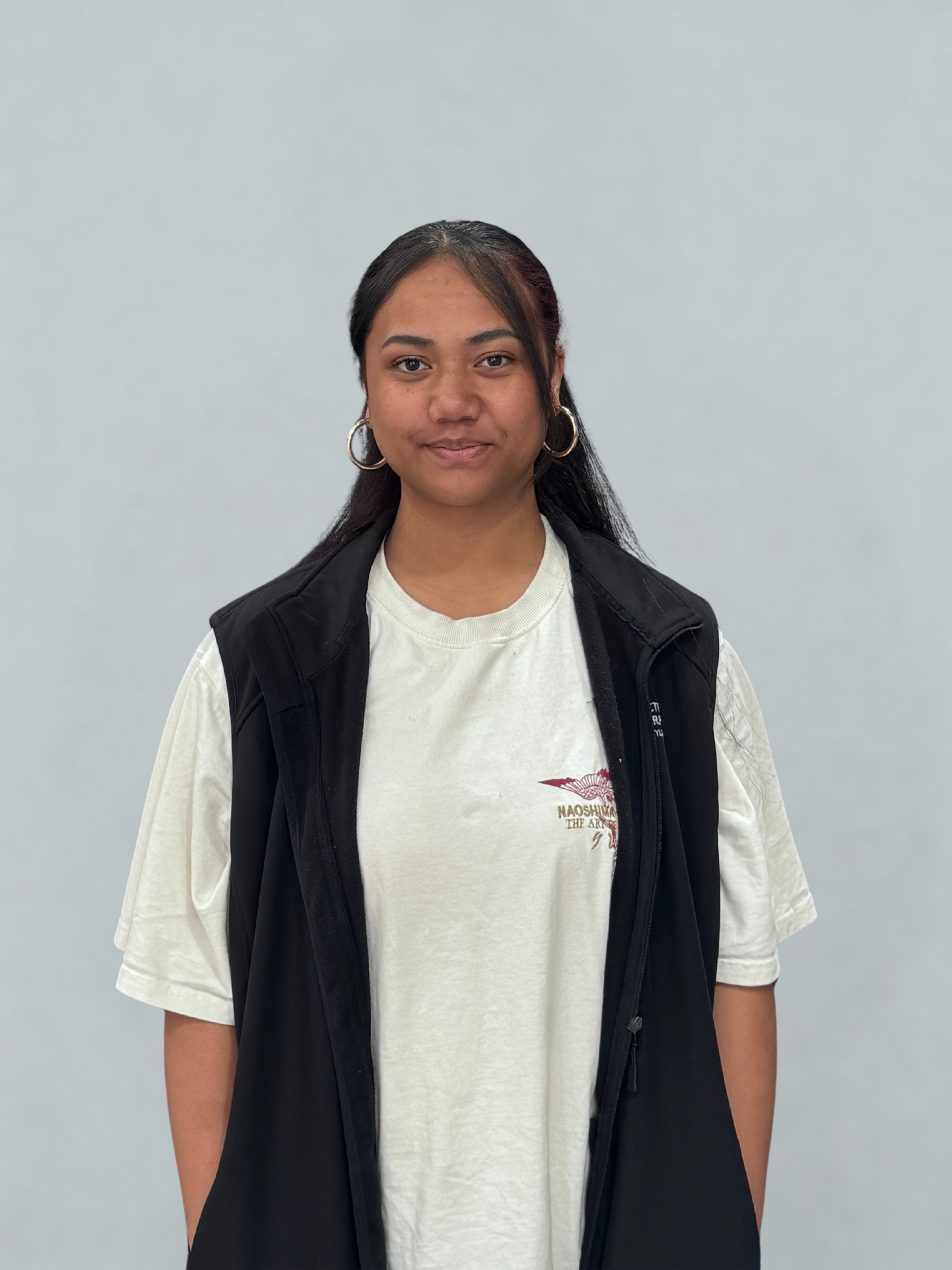 A young woman with medium skin tone, dark hair, wearing hoop earrings, a white t-shirt with a graphic logo, and a black vest, standing against a plain light gray background.