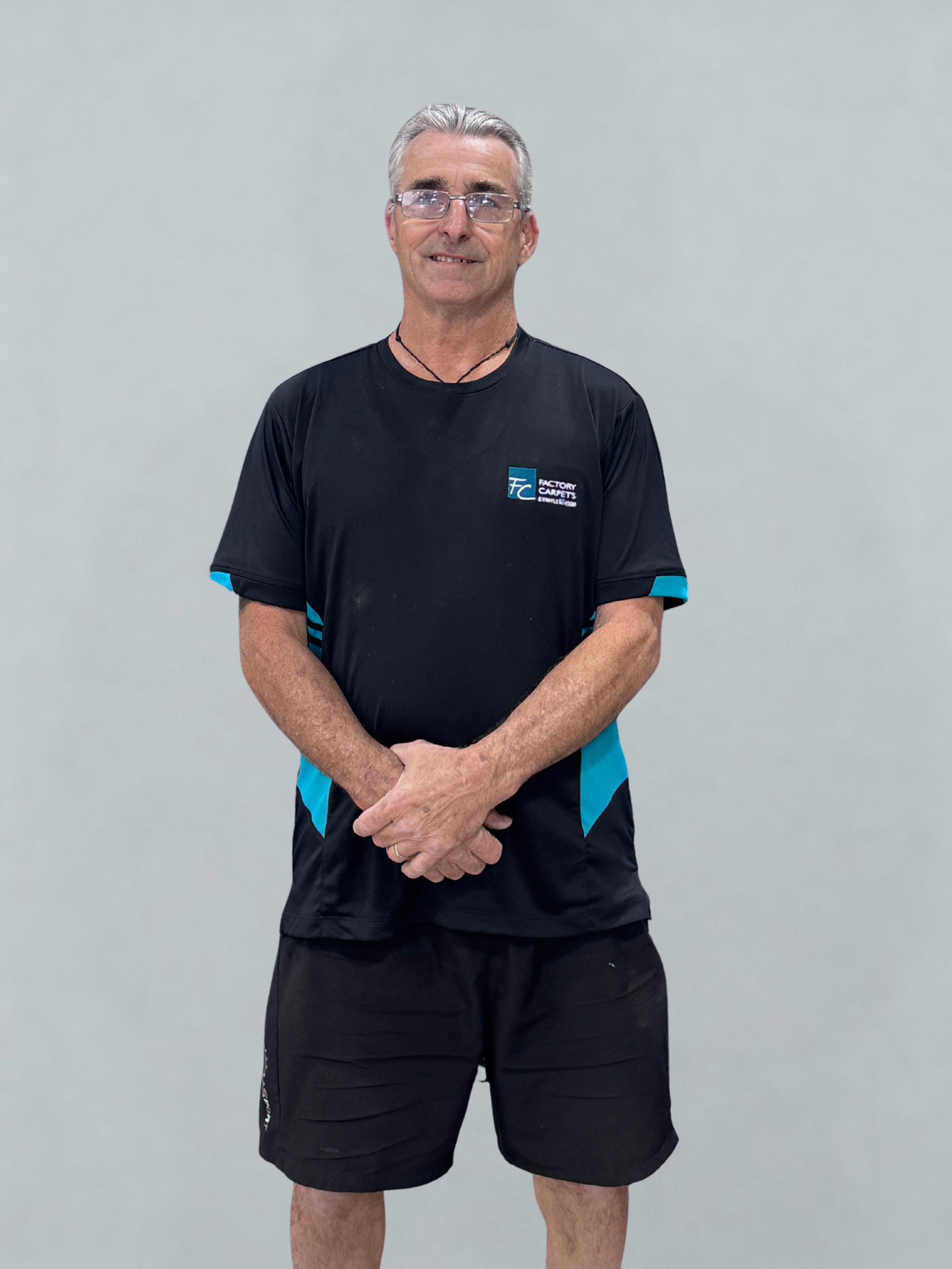 Man standing against plain background, wearing glasses, black and blue sports shirt, and black shorts, smiling.