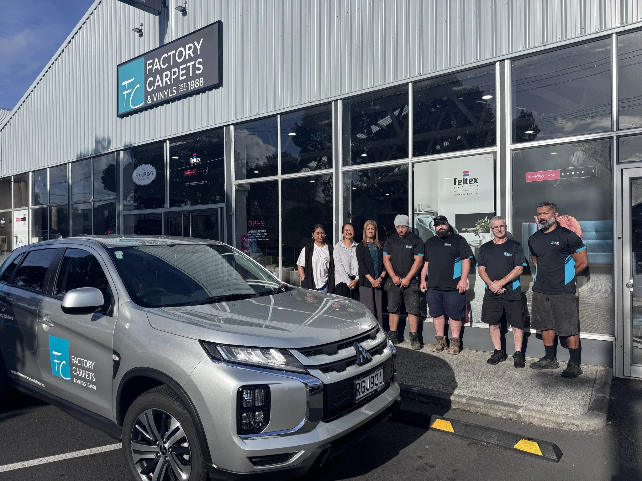 A group of seven people standing in front of a storefront with large glass windows and a sign that reads 'Factory Carpets & Vinyls Est. 1988'. There is a silver vehicle parked in front of the store with a logo and text that reads 'Factory Carpets & Vinyls'. The store appears to be a carpet and flooring business.