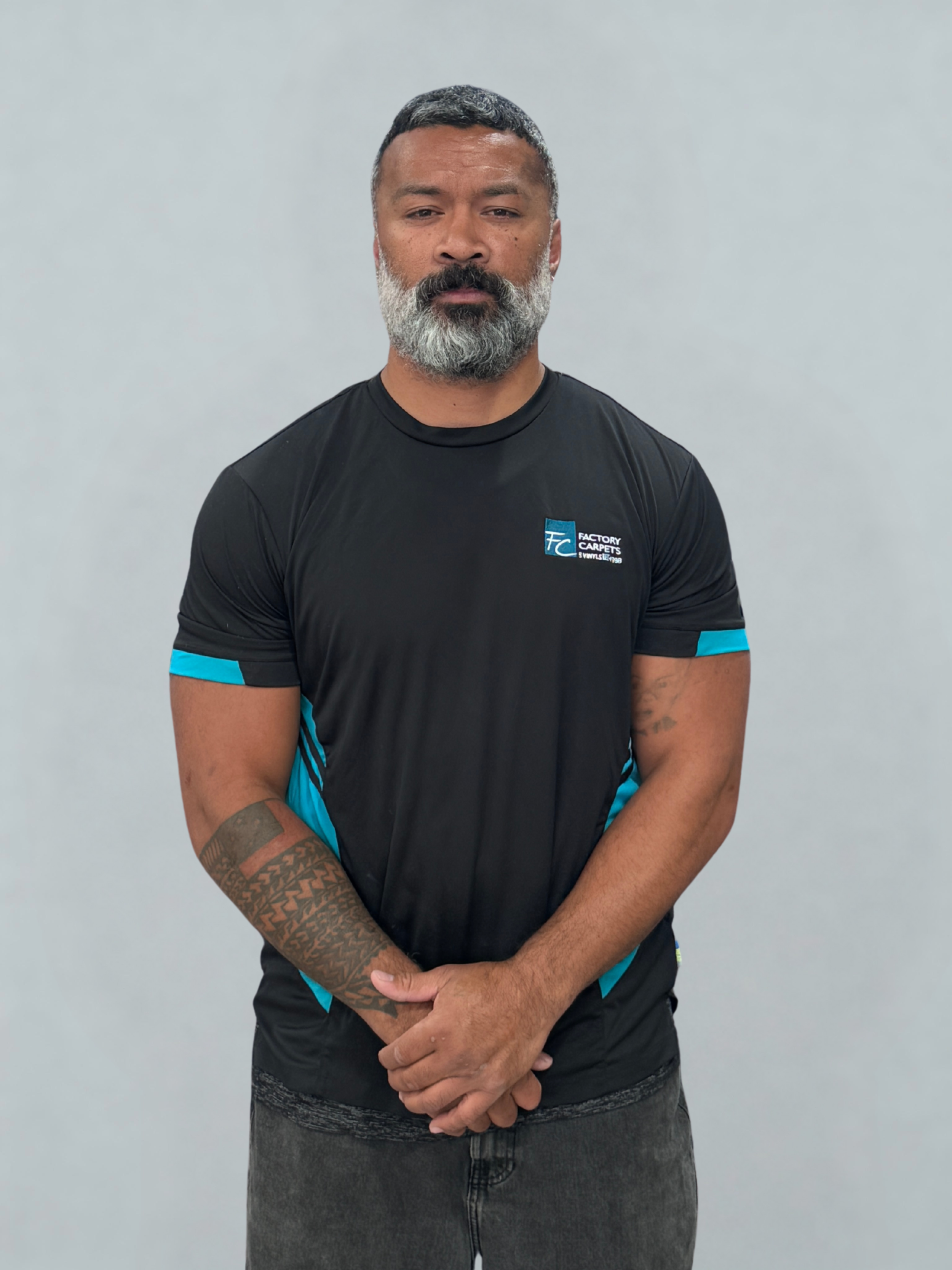 A man with a gray beard and short dark hair standing with his hands clasped in front of him, wearing a black and blue T-shirt with a logo that reads "Factory Careers" on the chest, against a plain light gray background.