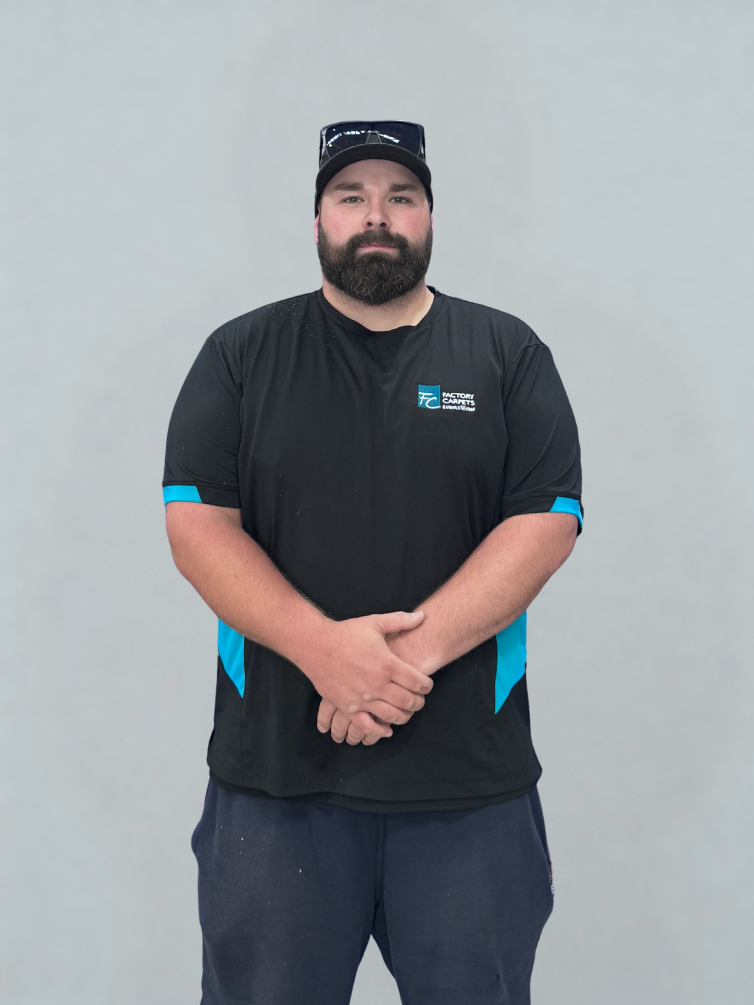 A man with a beard wearing a black Factory Carpets shirt, black cap, sunglasses, and navy pants standing against a plain gray background.