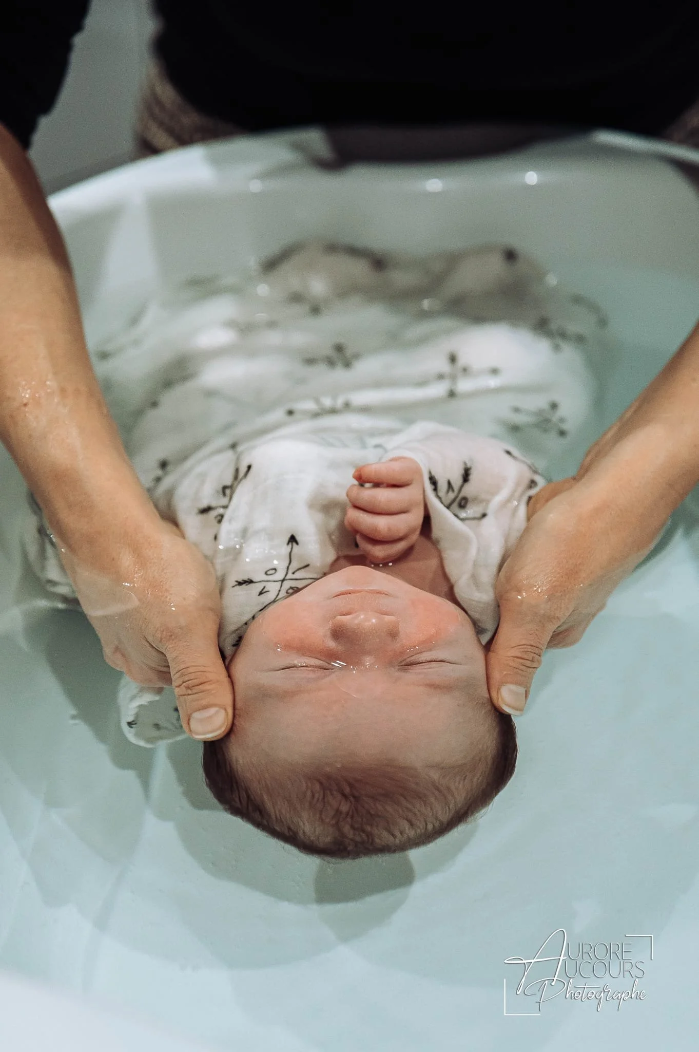 Bébé prenant un bain enveloppé dans un lange et bercé dans une baignoire par des mains d'adultes. on appelle ça un bain thérapeutique bébé méthode Sonia Krief, ou bain enveloppé.