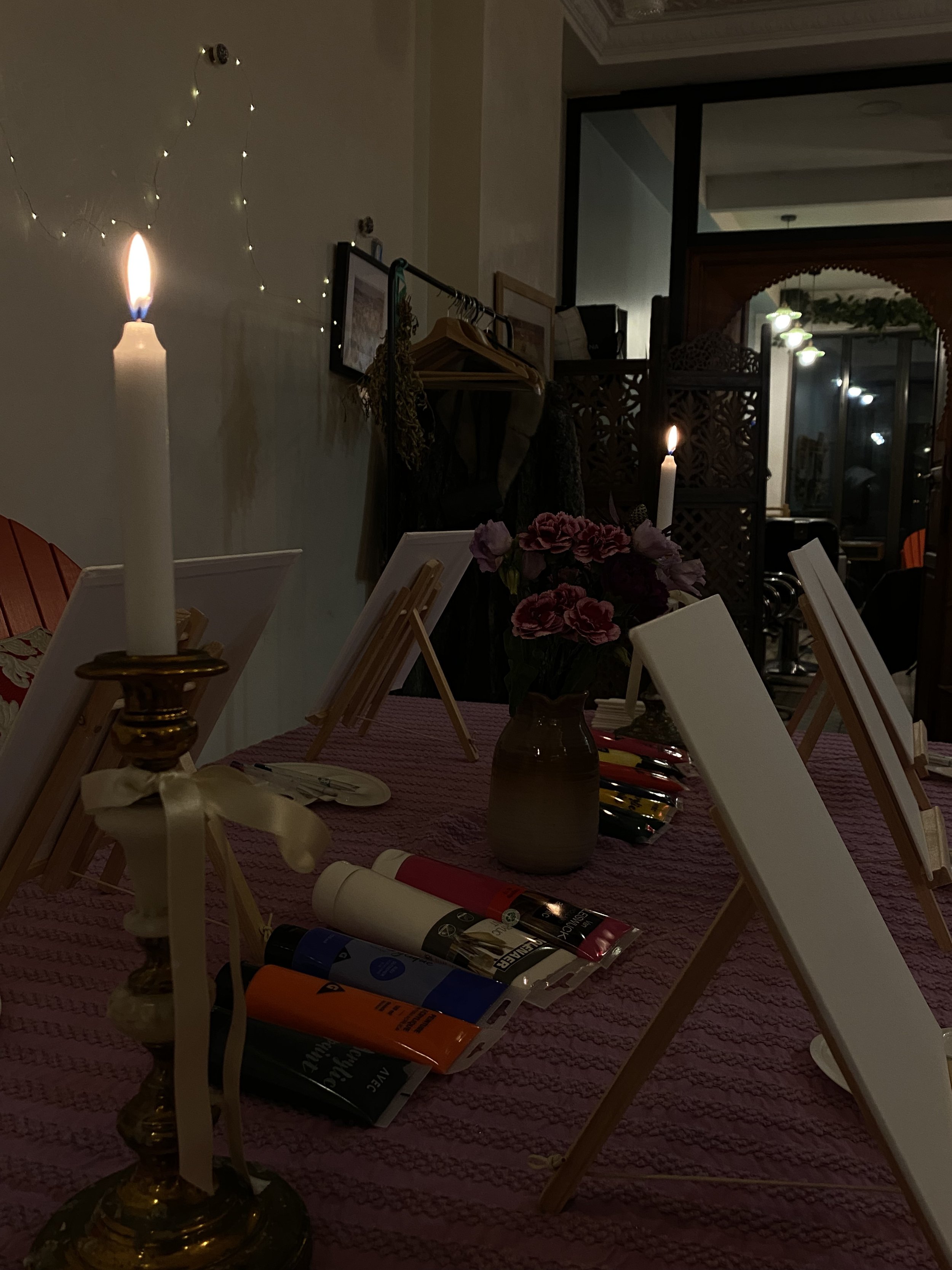 Table préparée pour une activité artistique avec chevalets, pots de peinture, chandeliers allumés et vase de fleurs.