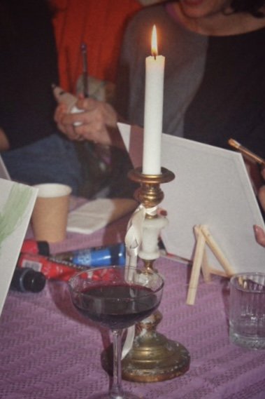 Une scène intérieure avec une table couverte d'une nappe violette. Sur la table, il y a un chandelier doré avec une bougie allumée, un verre de vin rouge, des pinceaux, des toiles blanches, et quelques tubes de peinture. Des personnes sont autour de