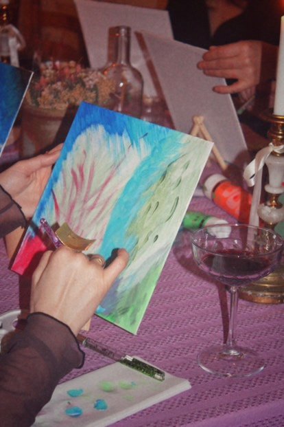 Personnes peignant à une table avec divers matériaux artistiques, y compris de la peinture acrylique, un verre de vin et des pinceaux.
