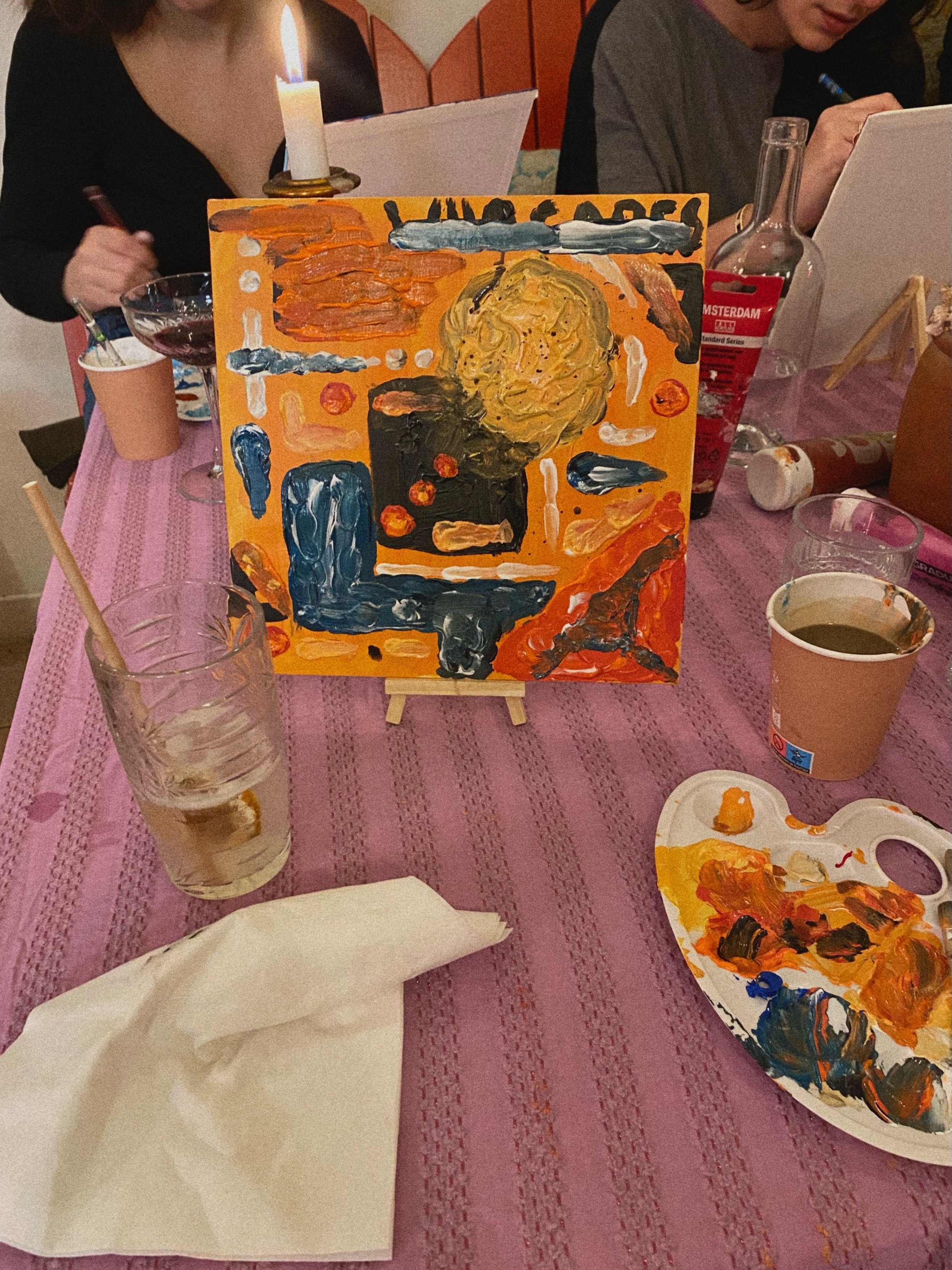 Scène d'une activité de peinture avec une toile colorée posée sur un chevalet. Des personnes peignent autour de la table, avec des palettes de peinture et divers outils. Un verre et une bouteille sont visibles, ainsi qu'une bougie allumée en arrière-