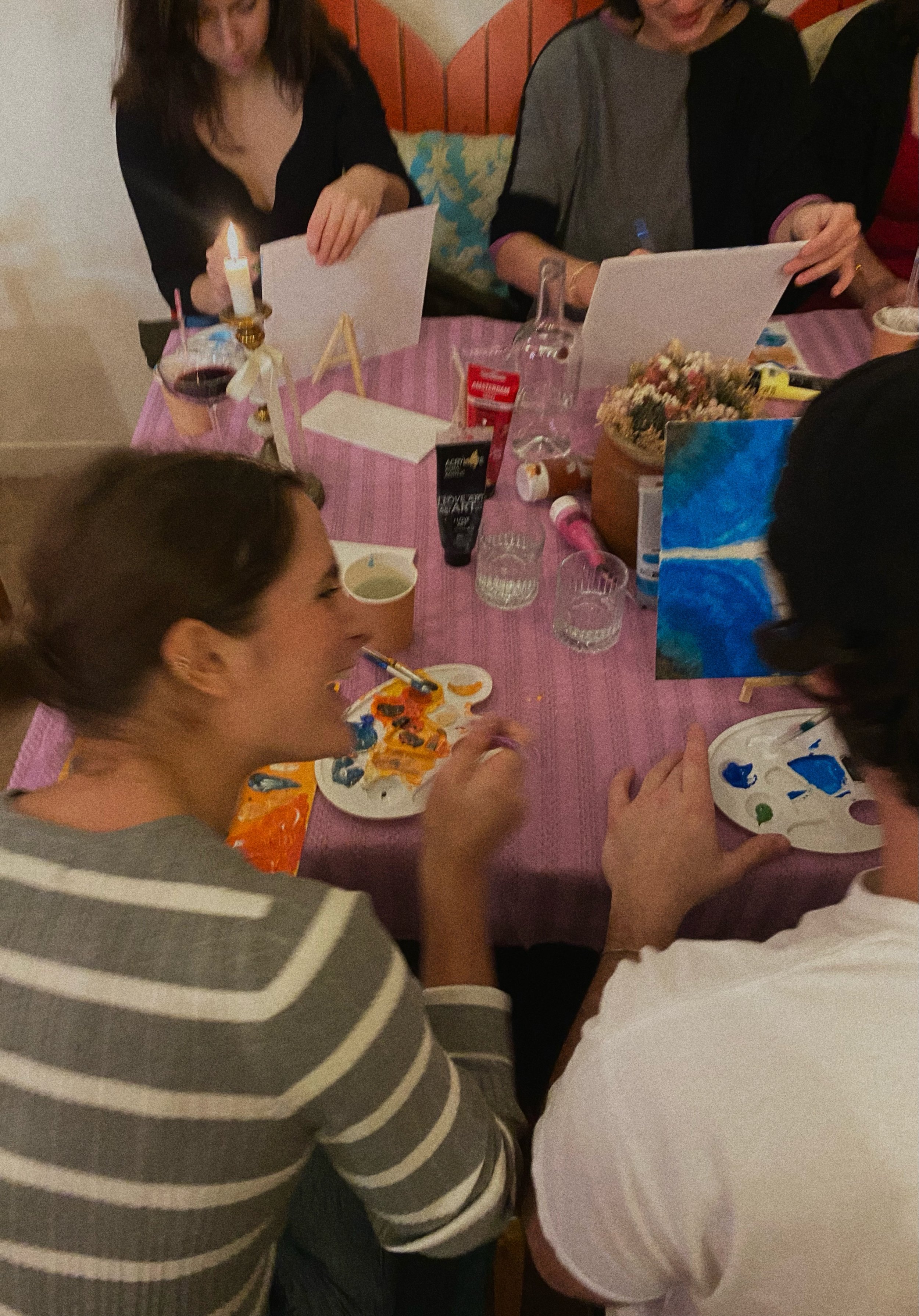 Des personnes autour d'une table peignant et faisant de l'artisanat sur une nappe violette avec des fournitures de peinture, des bougies et des boissons.