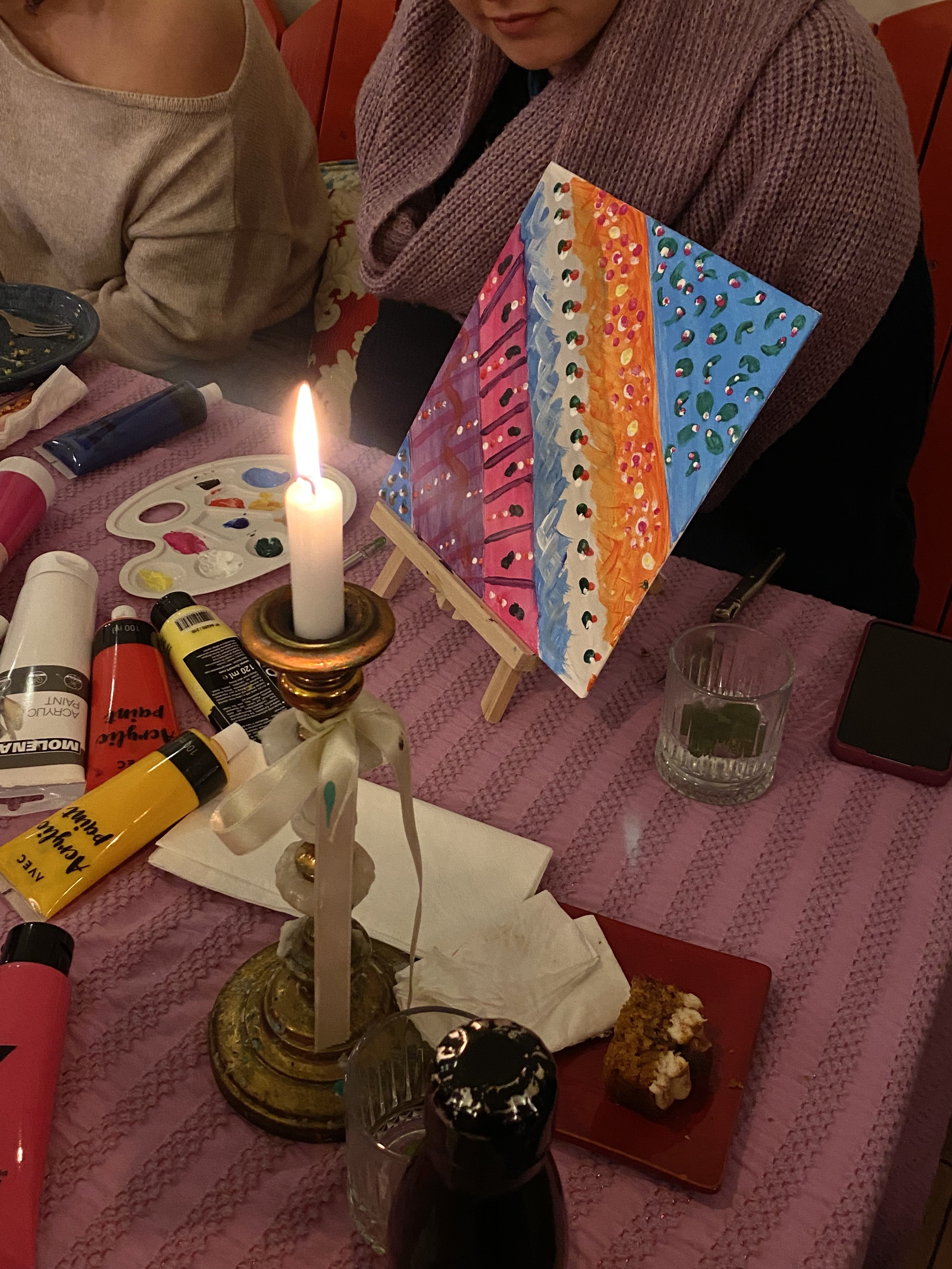 Deux personnes sont assises à une table décorée pour une activité artistique. Il y a des tubes de peinture acrylique, une palette de couleurs, une bougie allumée, un morceau de gâteau, et une toile peinte avec des motifs colorés sur un chevalet.