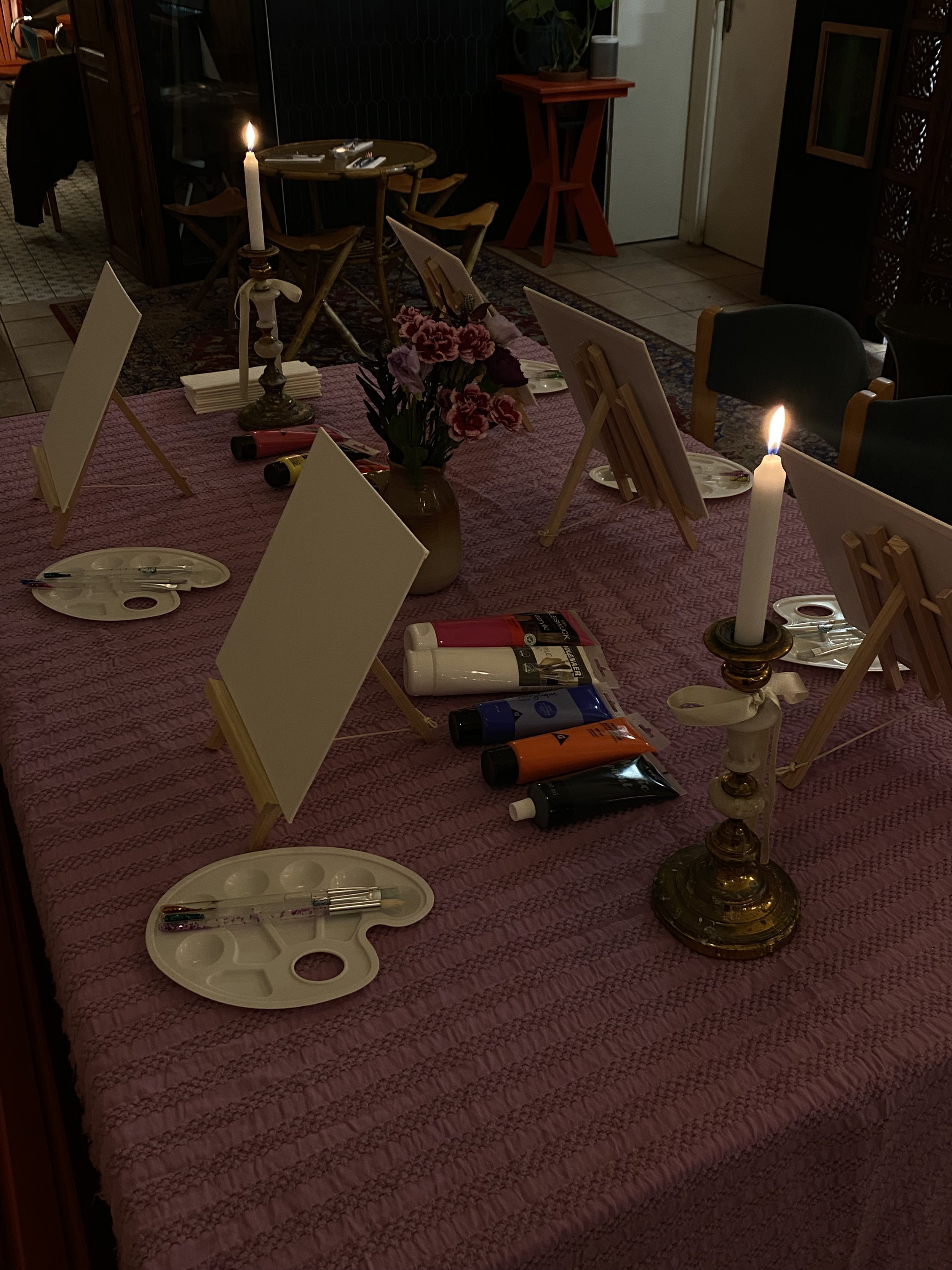 Table prévue pour une activité de peinture avec des chevalets, des toiles, palettes, pinceaux, tubes de peinture et bougies allumées, décorée avec des fleurs au centre.