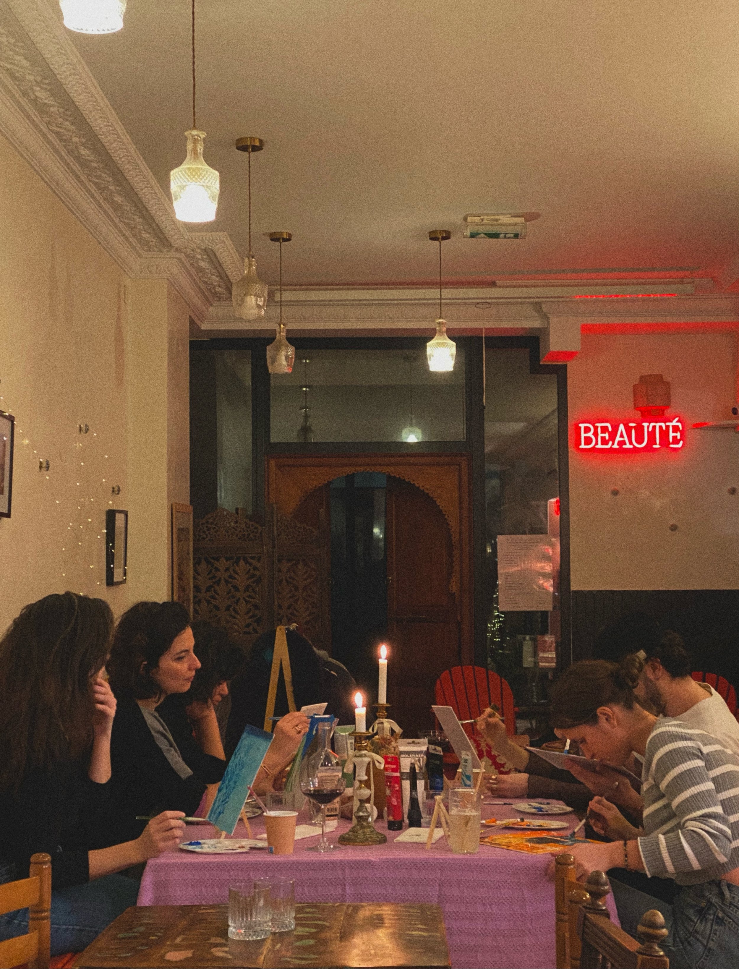 Un groupe de personnes assises autour d'une table lors d'une activité artistique dans un café. Elles peignent sur des toiles, avec des bougies allumées et des boissons sur la table. Un panneau lumineux "BEAUTÉ" est visible à l'arrière-plan.