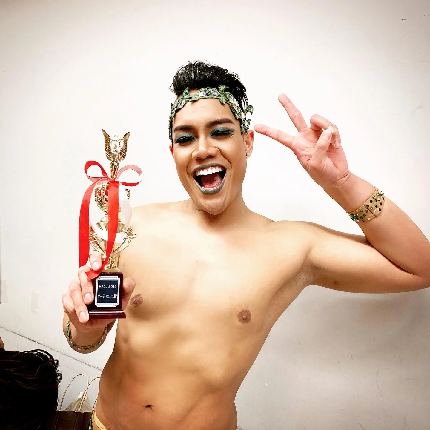 A shirtless person with short dark hair, makeup, and a decorative headband is holding a trophy in one hand and making a peace sign with the other, smiling widely.