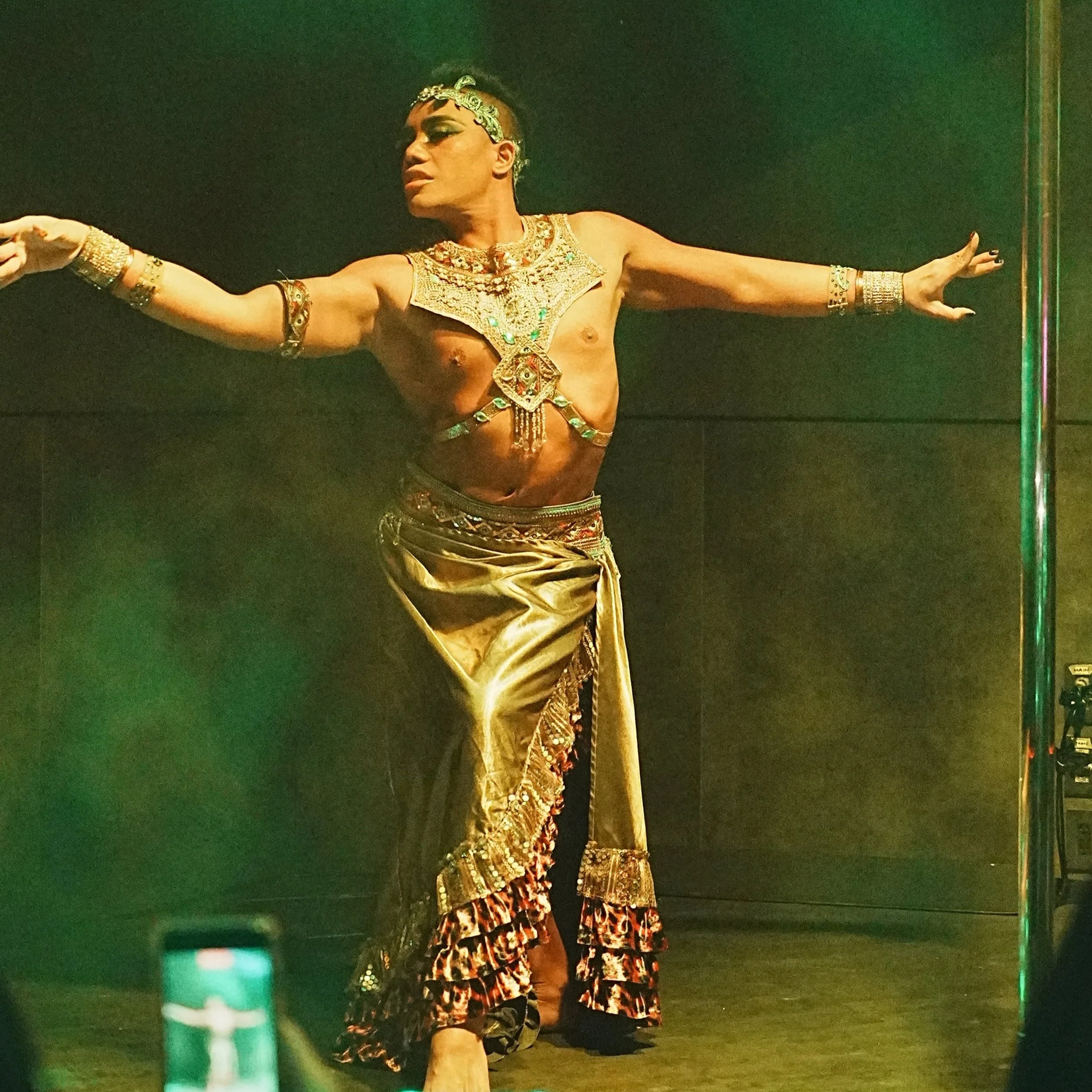 A performer dressed in elaborate gold costume with jewelry and a patterned skirt, dancing on stage with arms extended.