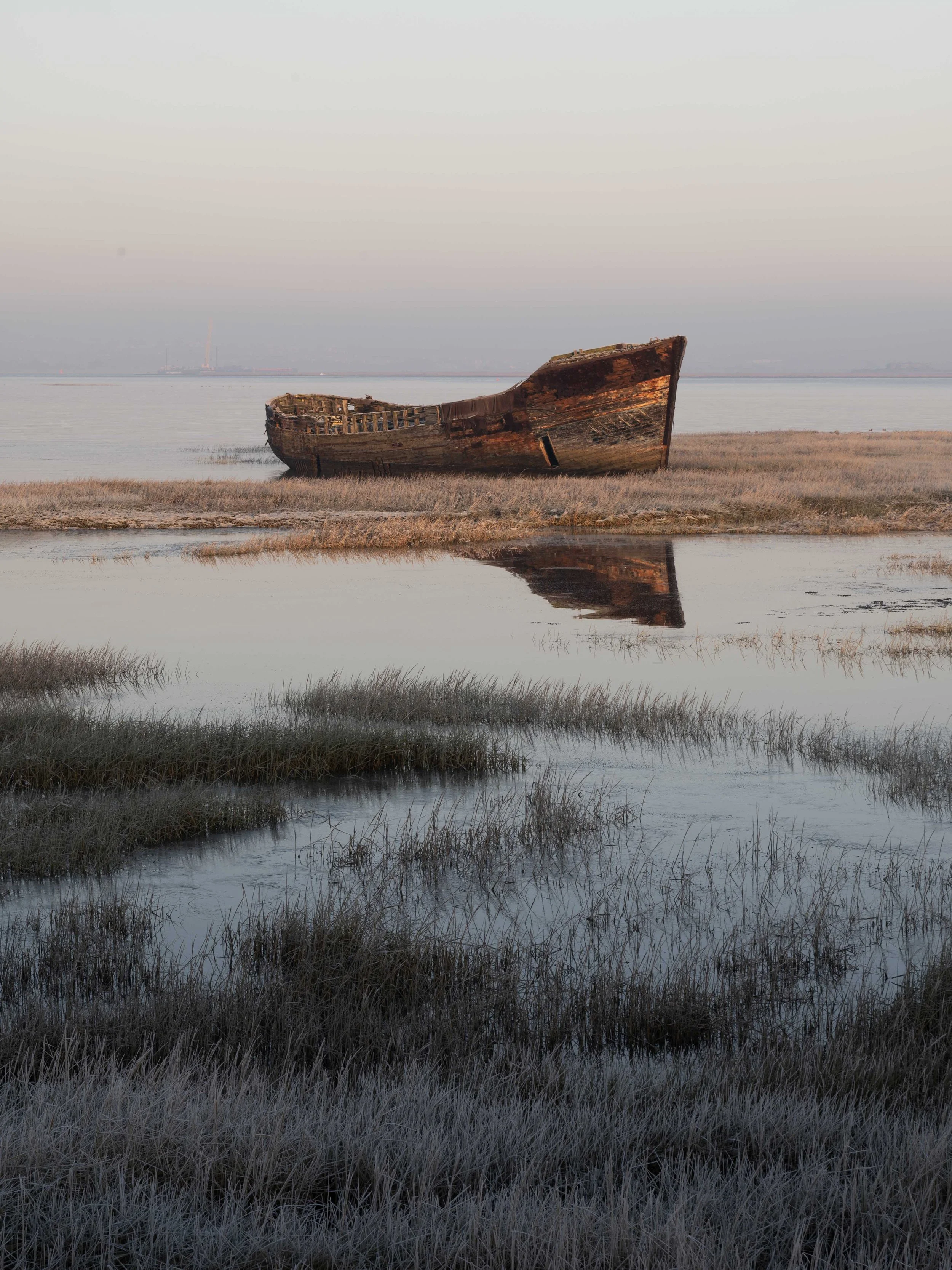 A rusted, abandoned boat resting on mud flats near water, with its reflection visible in the still water surface, under a pale sky.