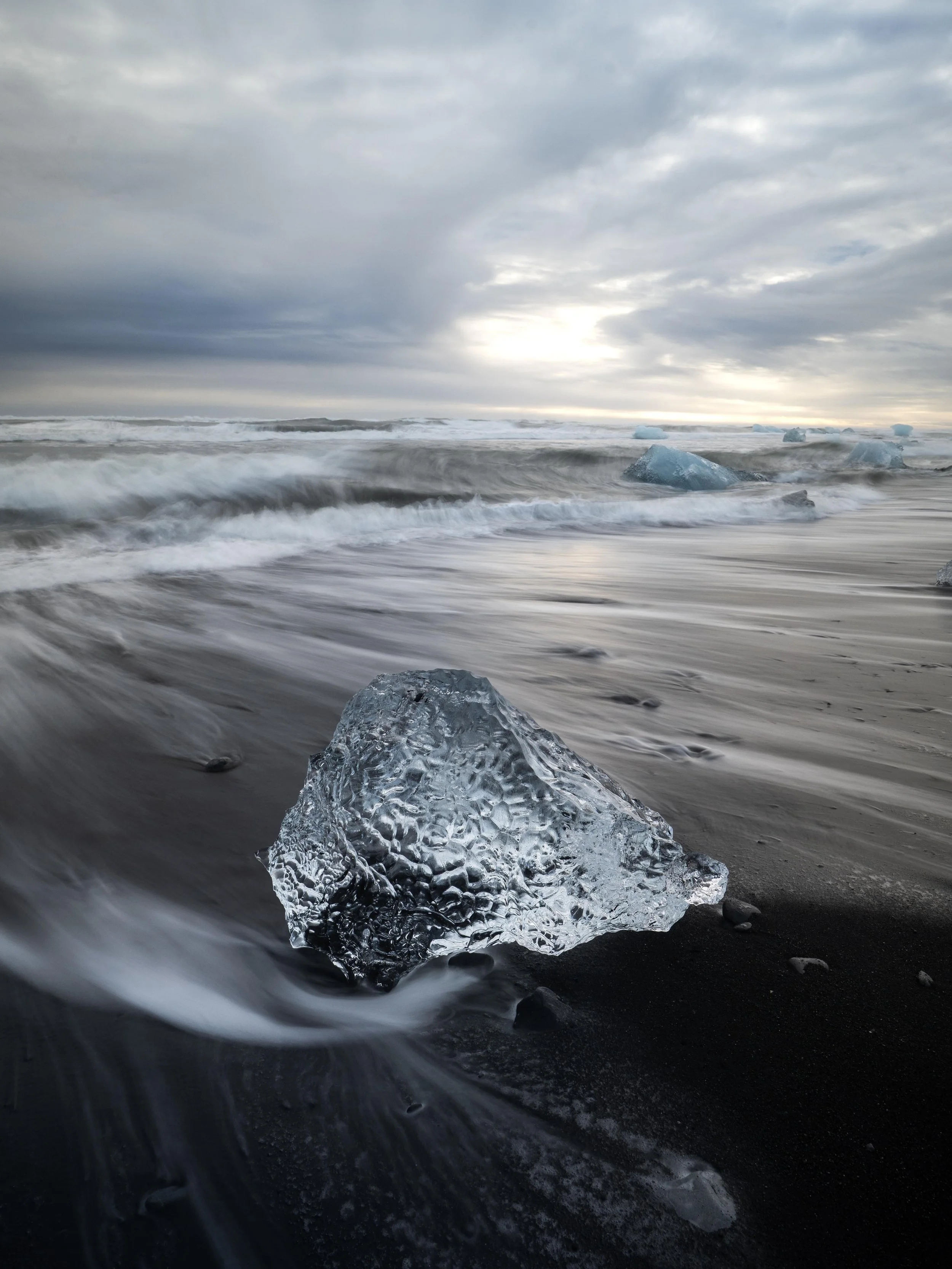 Iceberg on a black sand beach with waves crashing and a cloudy sky.